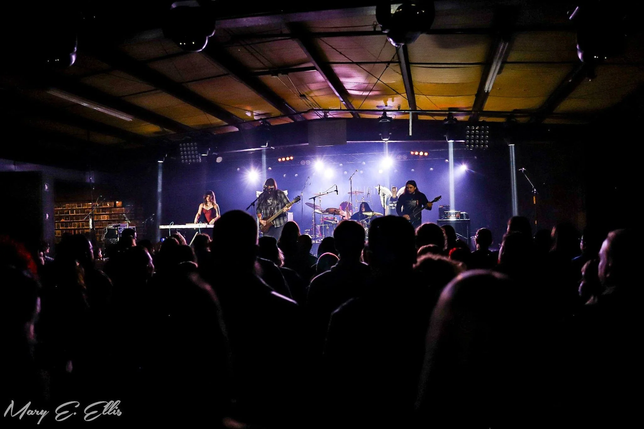 A band performing on stage at a concert venue with bright white and purple lighting, with audience members in the foreground.