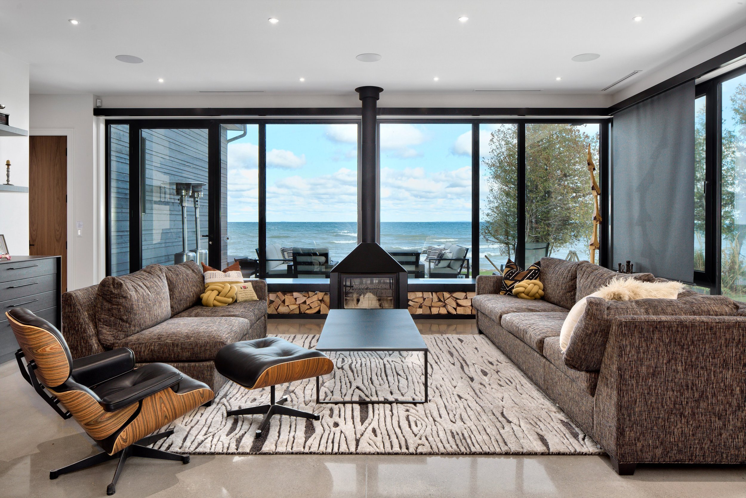 Mid century style living room with brown sofas, a black fireplace, large windows, and ocean view. Includes a wood and leather lounge chair with ottoman, a black coffee table, and patterned area rug.