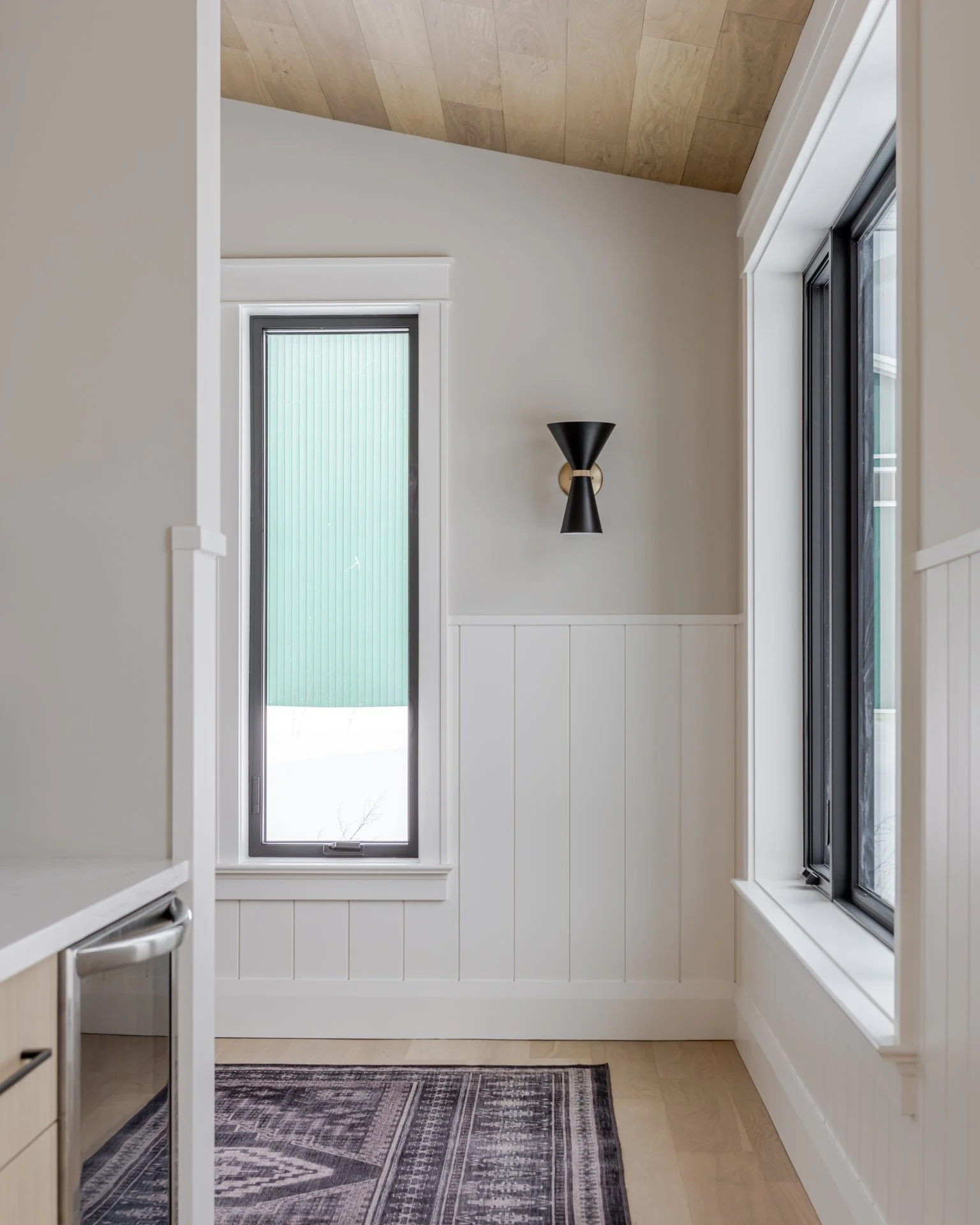 Modern hallway with a wooden ceiling, white panel walls, a black wall sconce, two vertical windows, and a patterned area rug on a wooden floor.