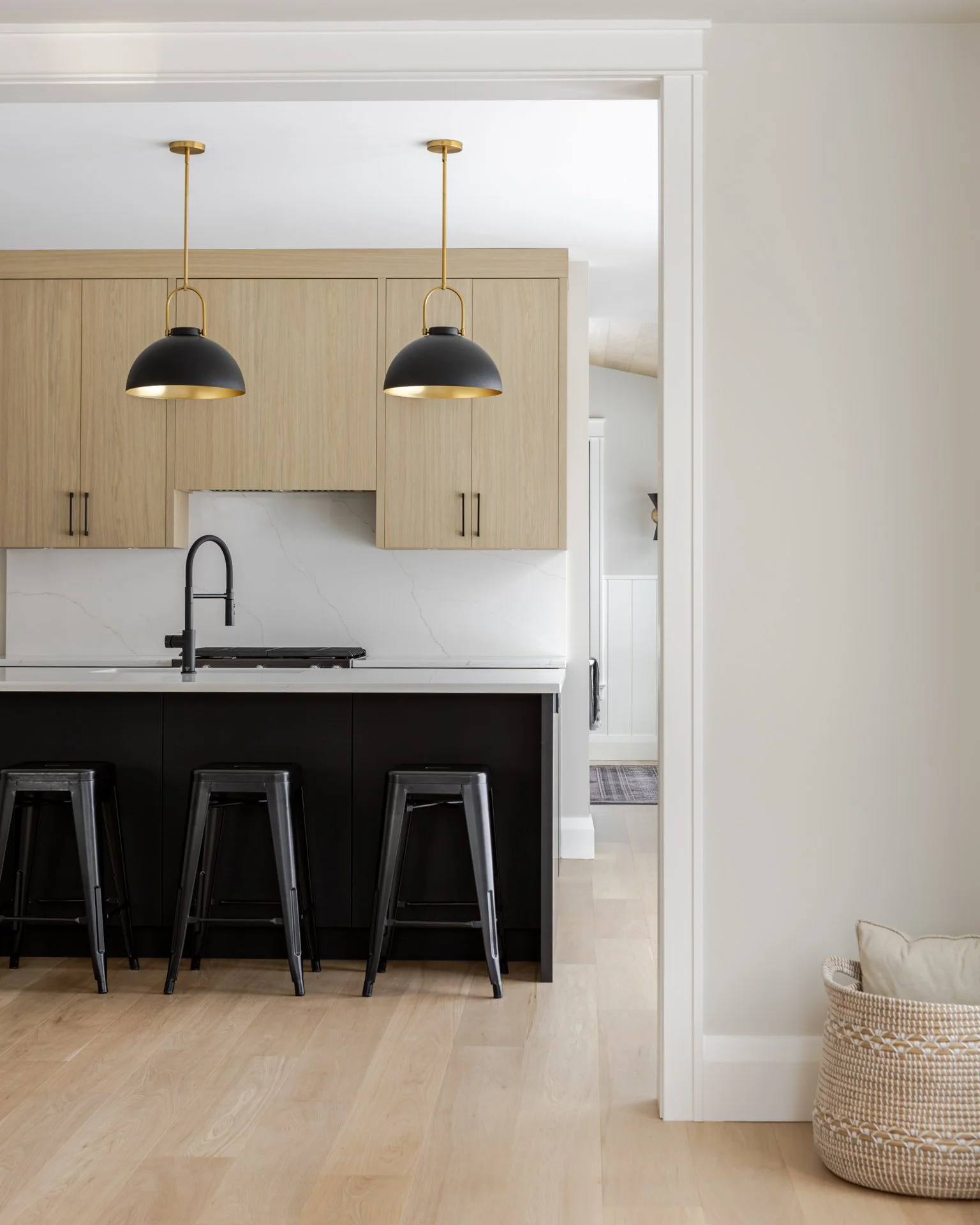Modern kitchen interior with light wooden cabinets, black kitchen island, and black bar stools. Two black and gold pendant lights hang above the island. A woven basket with a cushion is in the foreground.