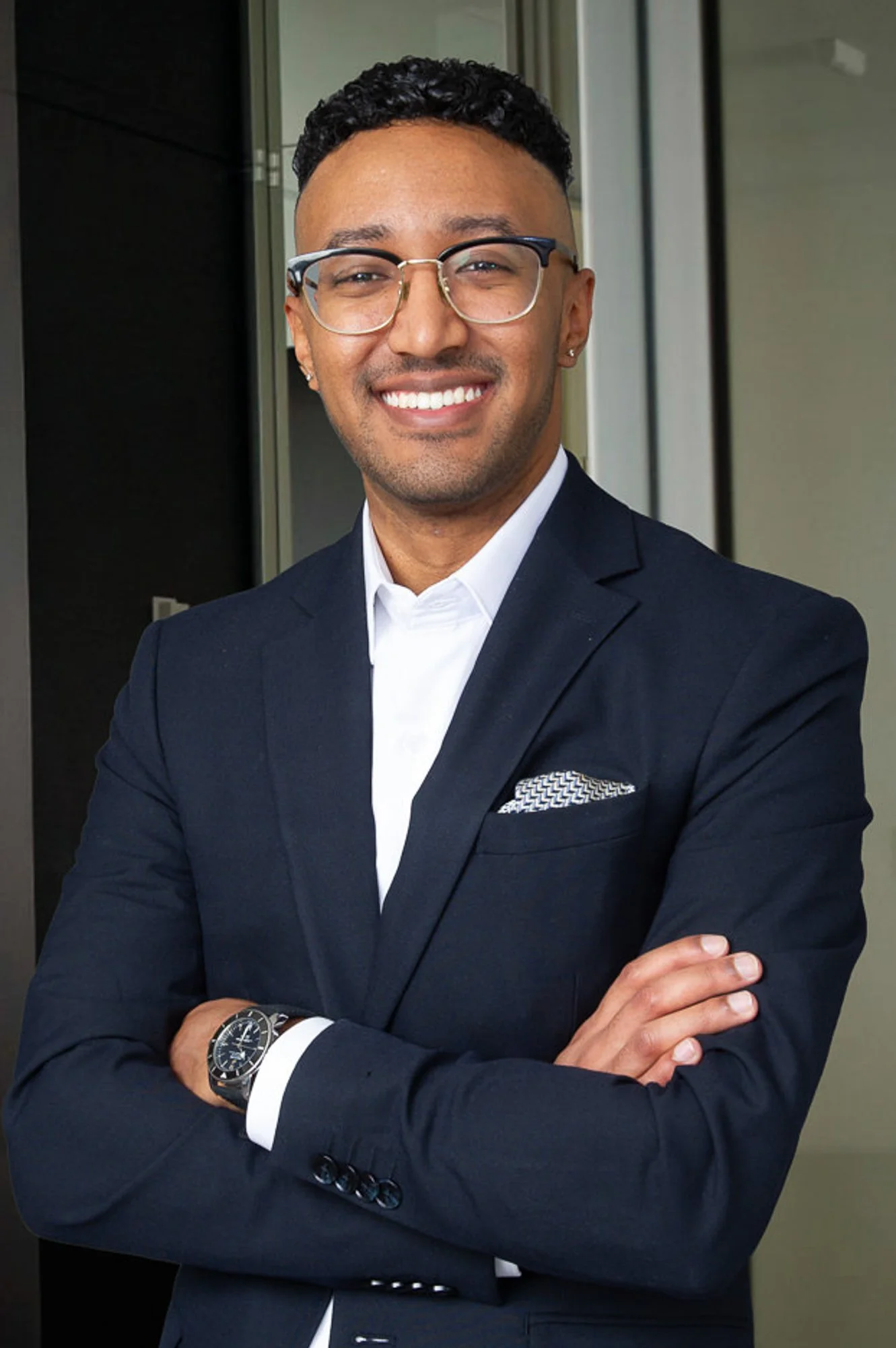 Man in formal attire with glasses smiling and standing with arms crossed.
