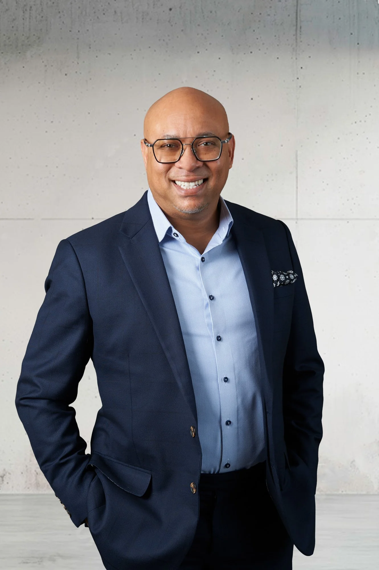Man wearing glasses and a navy suit with a light blue shirt, smiling against a light background.