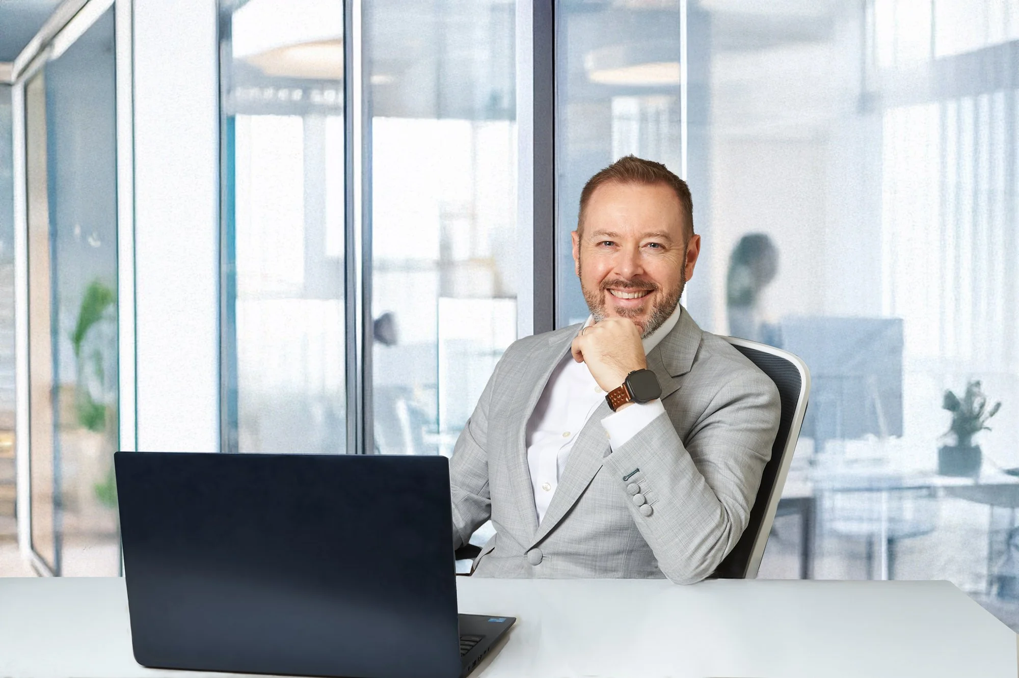 Man in gray suit smiling at a desk with a laptop in a modern office.