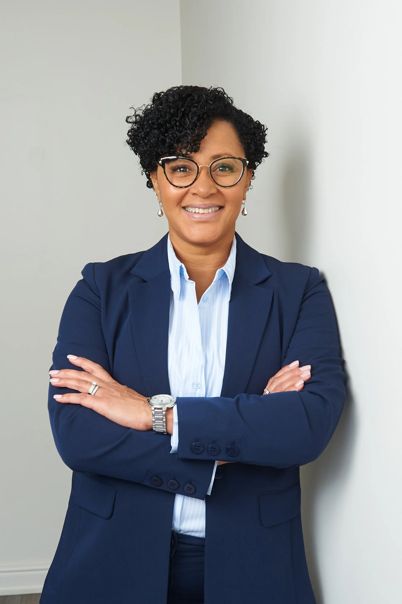 Person in a blue suit with crossed arms, smiling, wearing glasses and a watch, standing against a white wall.