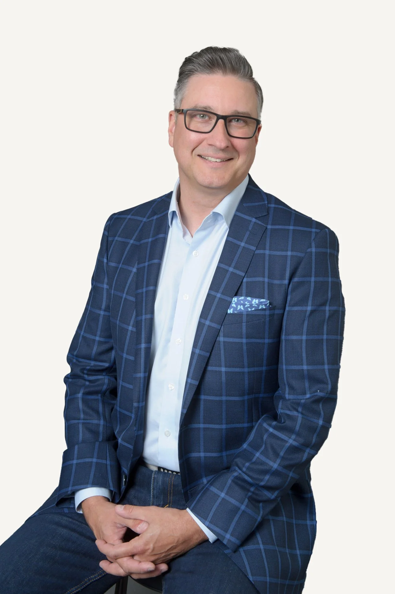 A man wearing glasses and a blue checkered blazer with a light blue shirt, sitting and smiling, against a light background.