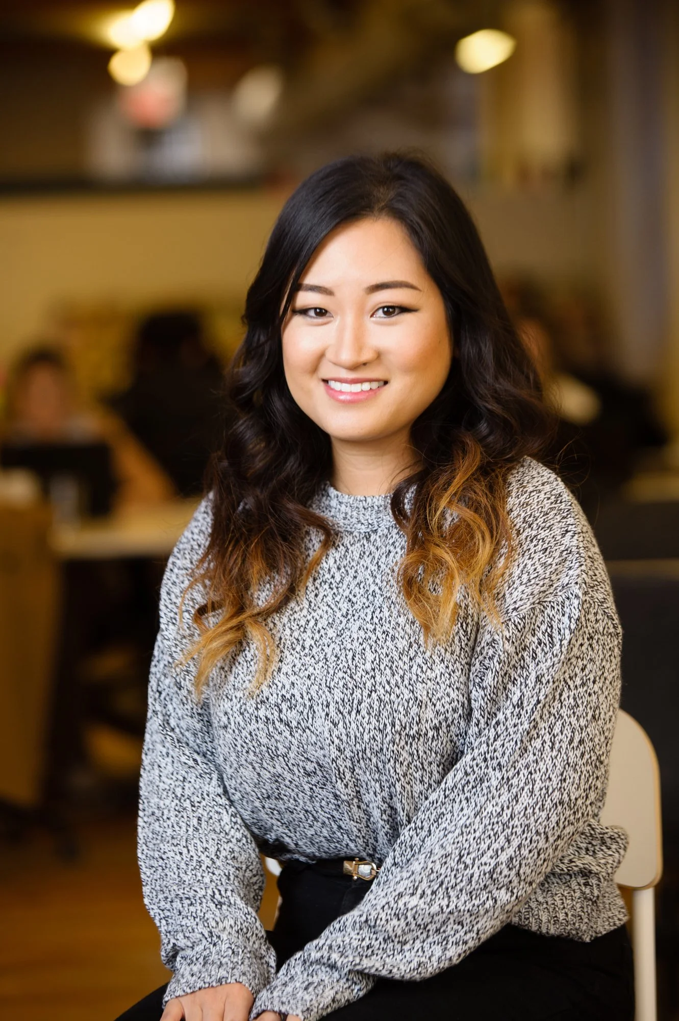 Woman with long hair wearing a gray sweater, smiling in an indoor setting.