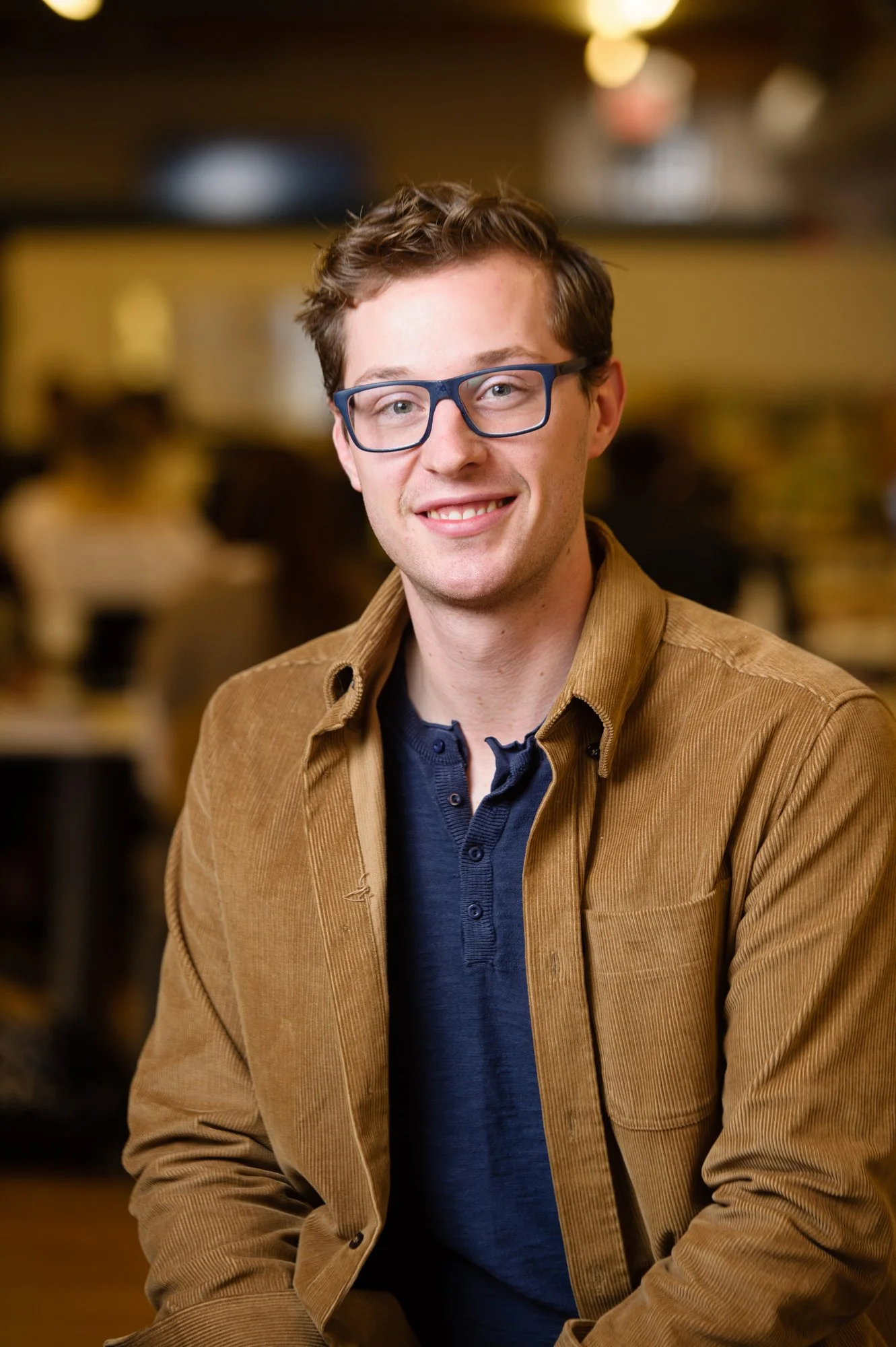 Man wearing glasses and a brown jacket, smiling indoors.