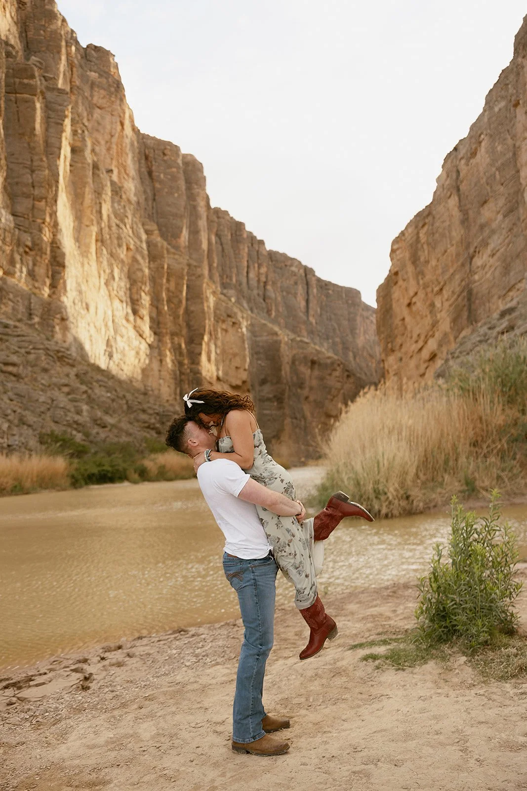 A couple embracing, with the woman lifted off the ground, standing near a river with tall canyon walls in the background.