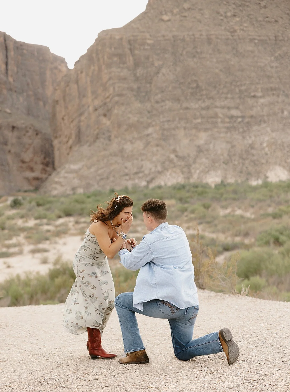 A man proposing to a woman in a desert landscape with mountains in the background. The woman is visibly surprised and emotional, covering her mouth with her hand, while the man is kneeling on one knee holding her hands.