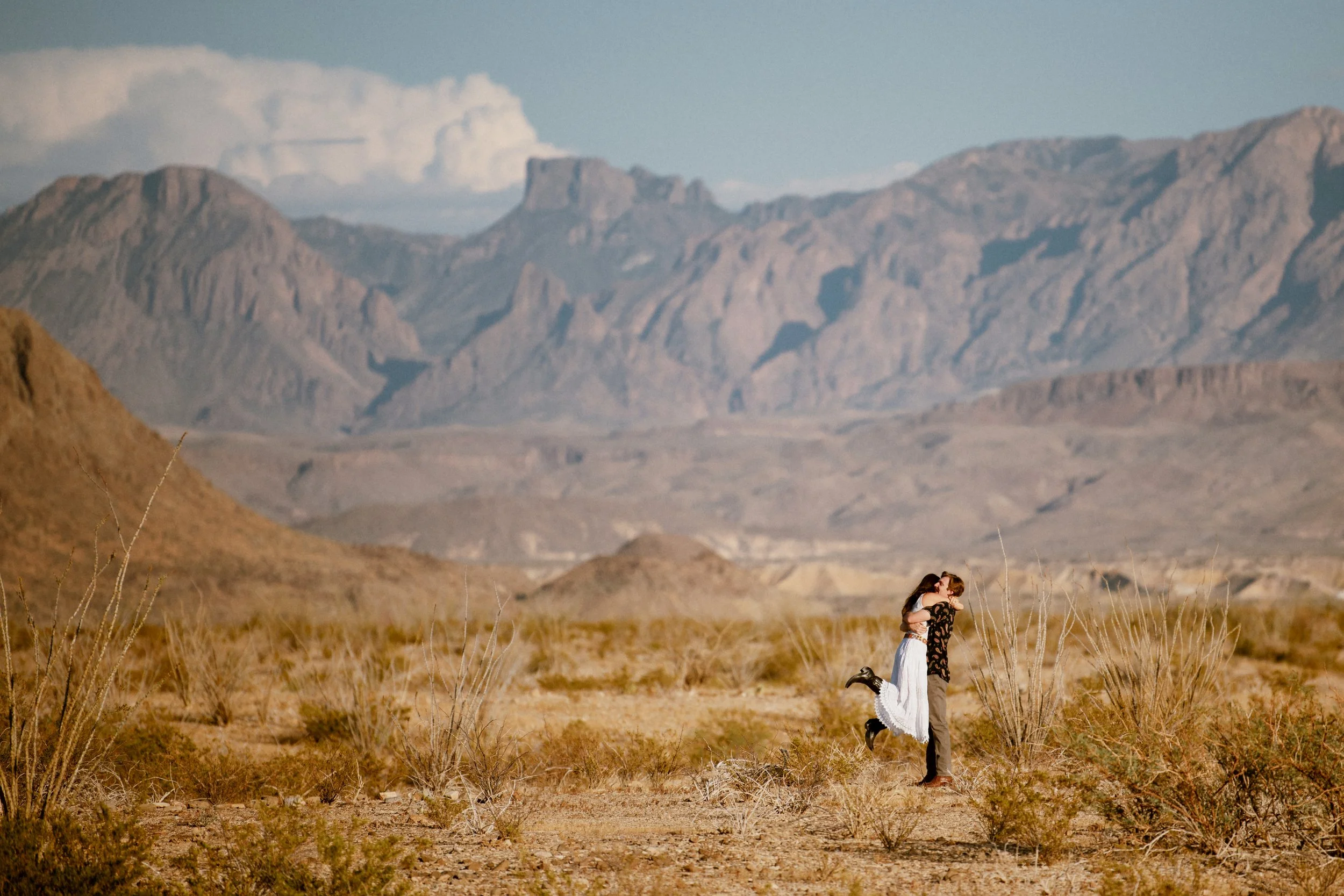 A couple embraces in a desert landscape with mountains in the background.