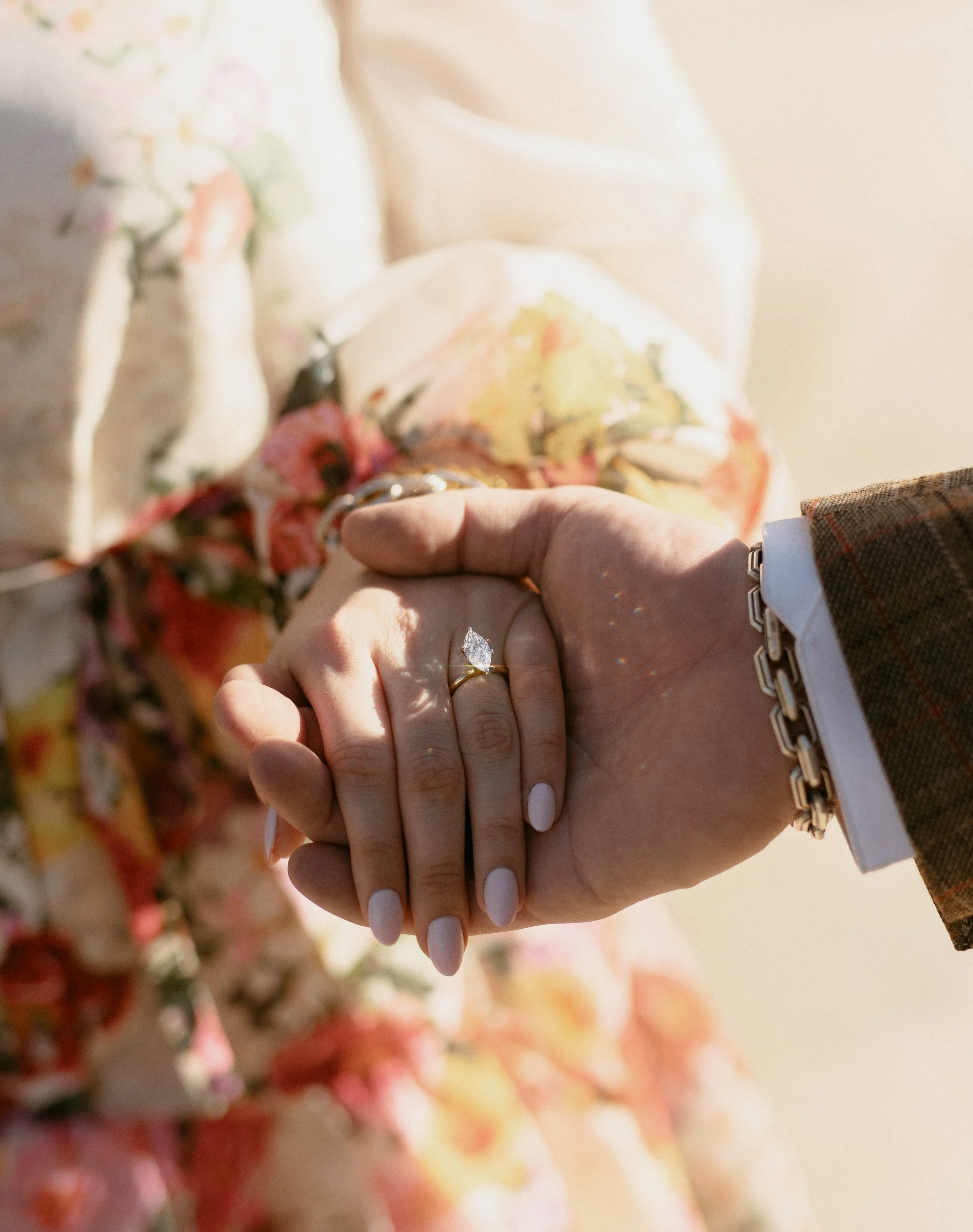 Close-up of a couple holding hands, showing an engagement ring on the woman's finger. The woman is wearing a floral dress, and the man is wearing a suit with a plaid sleeve and a bracelet. The background is blurred with warm lighting.