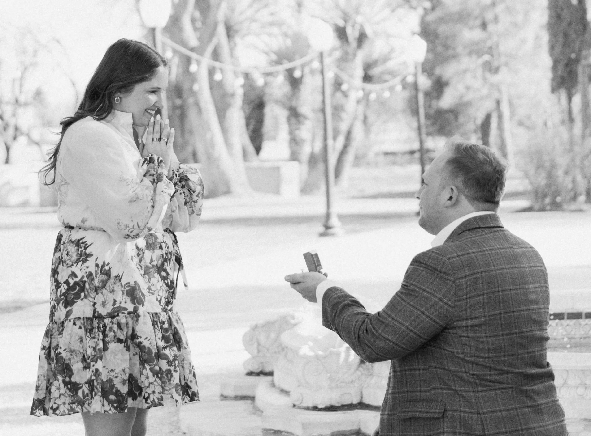 A man on one knee holding a ring in front of a woman who appears to be surprised and happy, in an outdoor park setting with trees and string lights in the background.