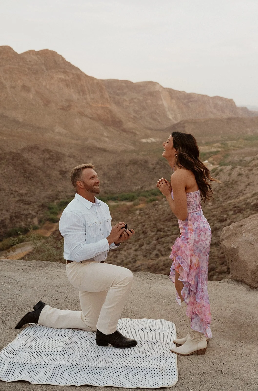 A man proposing marriage to a woman in a rocky desert landscape, with mountains in the background during sunset.