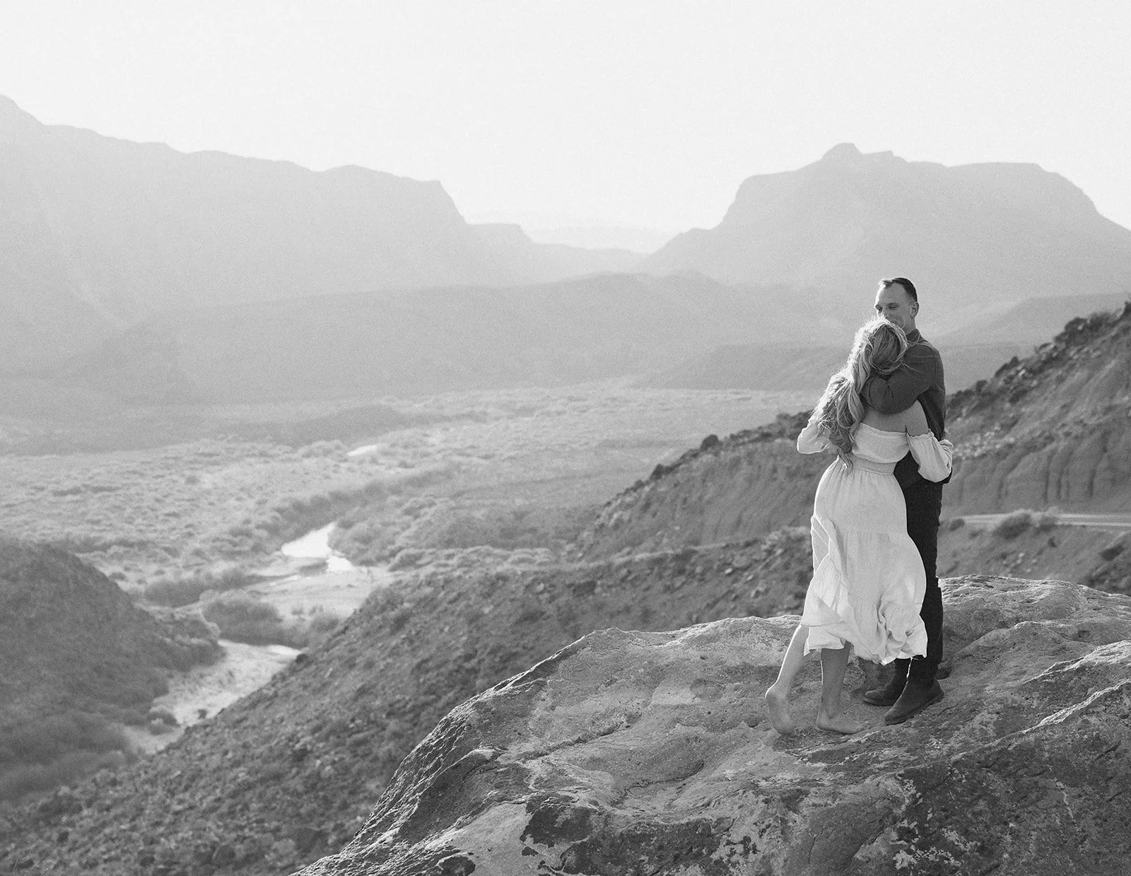 A black and white photo of a couple embracing on a rocky outcrop in a mountainous landscape, with a river winding through the valley below.