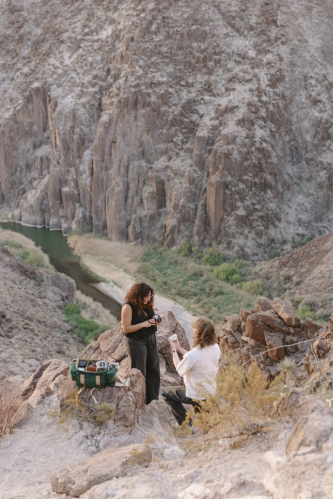 Two women in a desert canyon, one standing and holding a camera, the other sitting and holding a piece of paper, with a green cooler nearby.