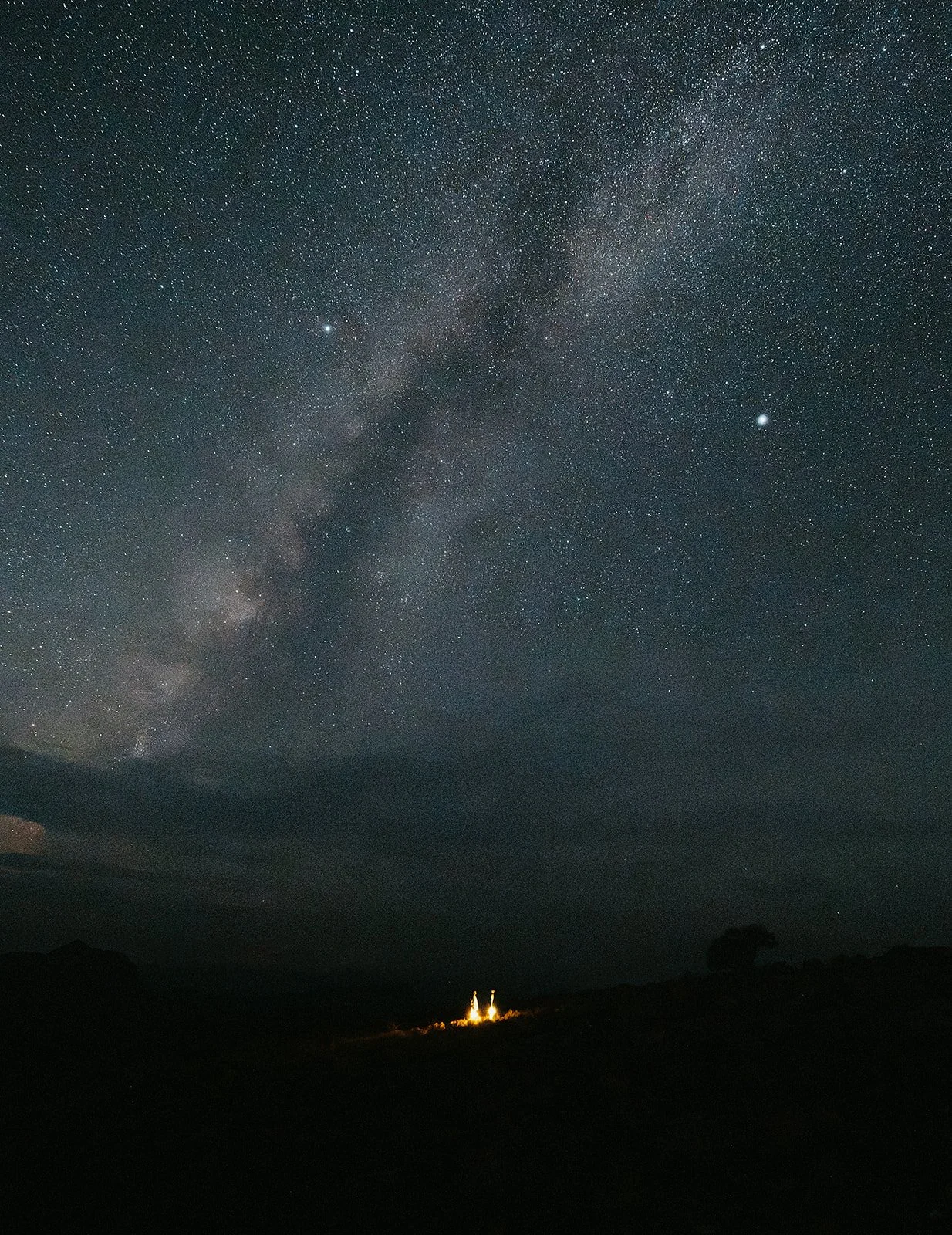 Night sky filled with numerous stars and the Milky Way galaxy, with a small illuminated structure or house on the horizon.