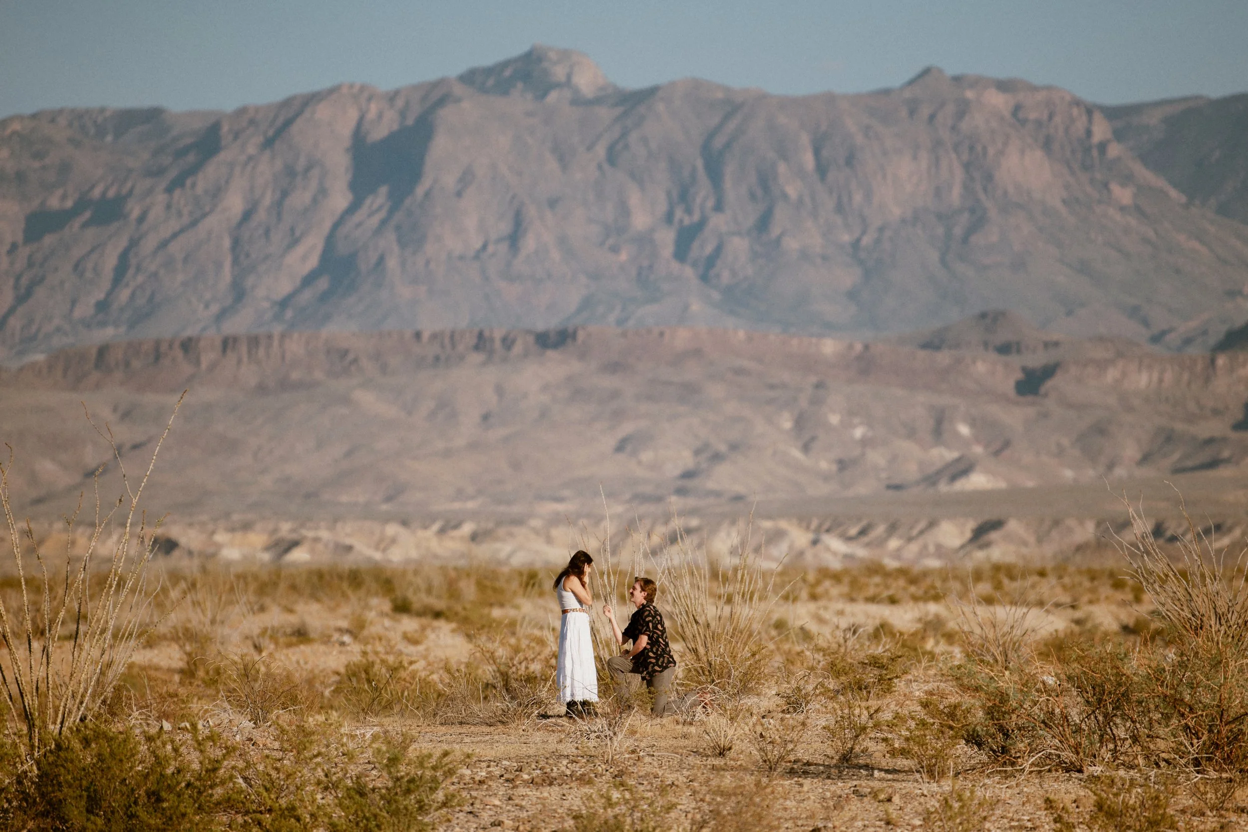 A man proposing to a woman in a desert landscape with mountains in the background.