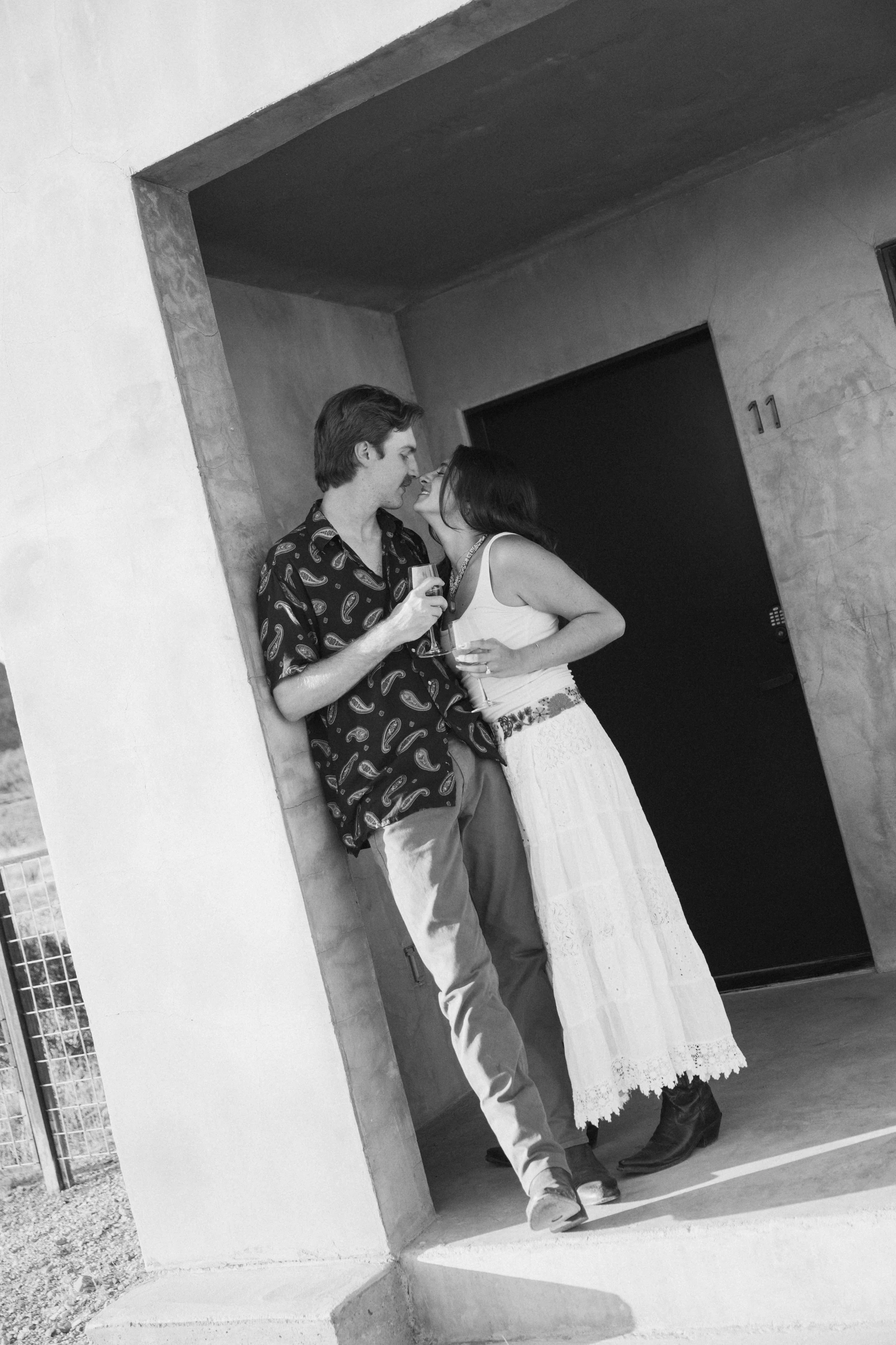 A black and white photo of a young man and woman standing close together outside, leaning against a wall, sharing drinks, and looking at each other affectionately.