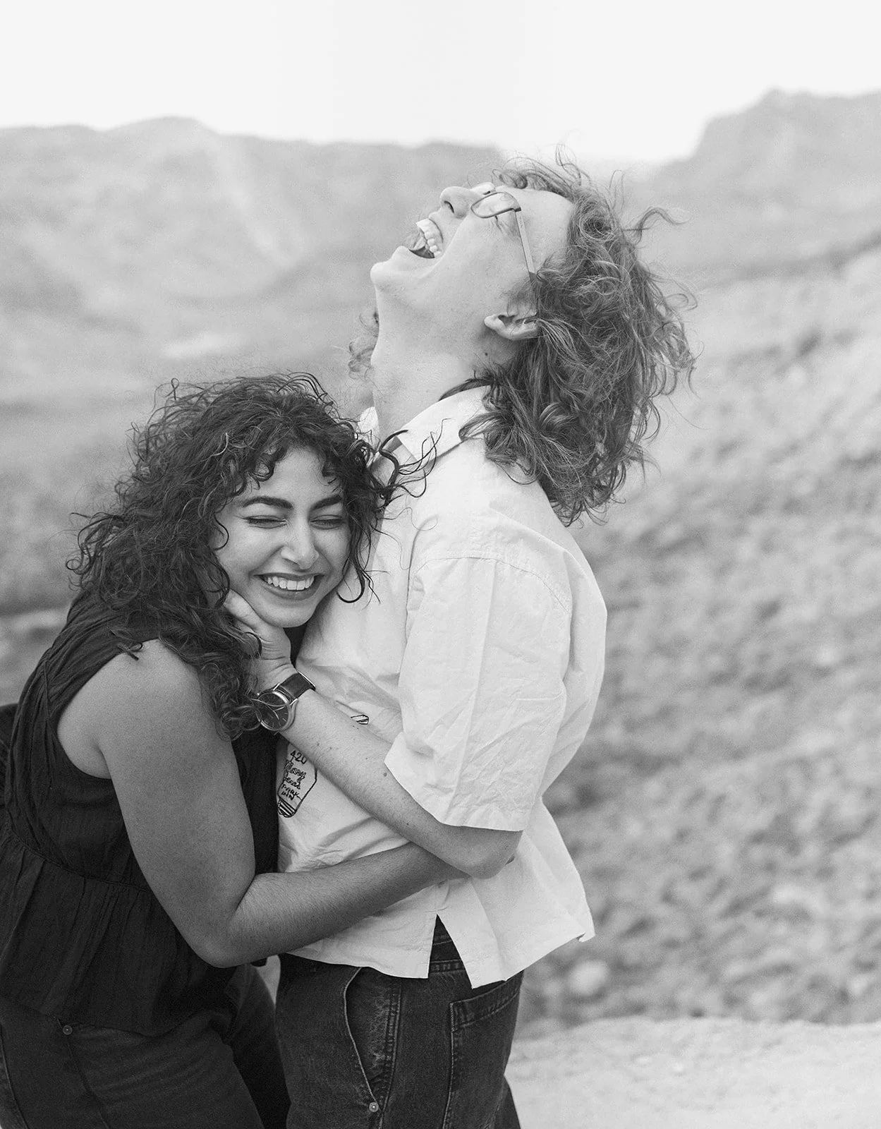Two women with curly hair laughing and hugging outdoors with mountains in the background, black and white photo.