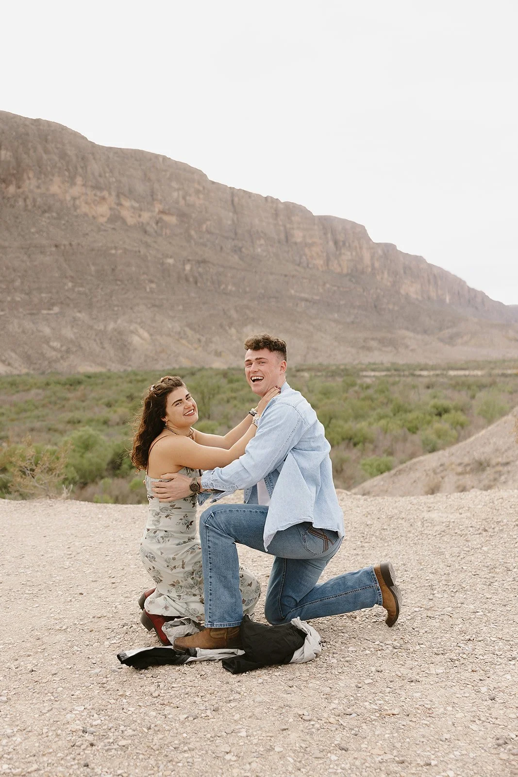 A young man proposing marriage to a woman in a desert landscape with mountains in the background. The woman is smiling and touching the man's face, who is kneeling on one knee.