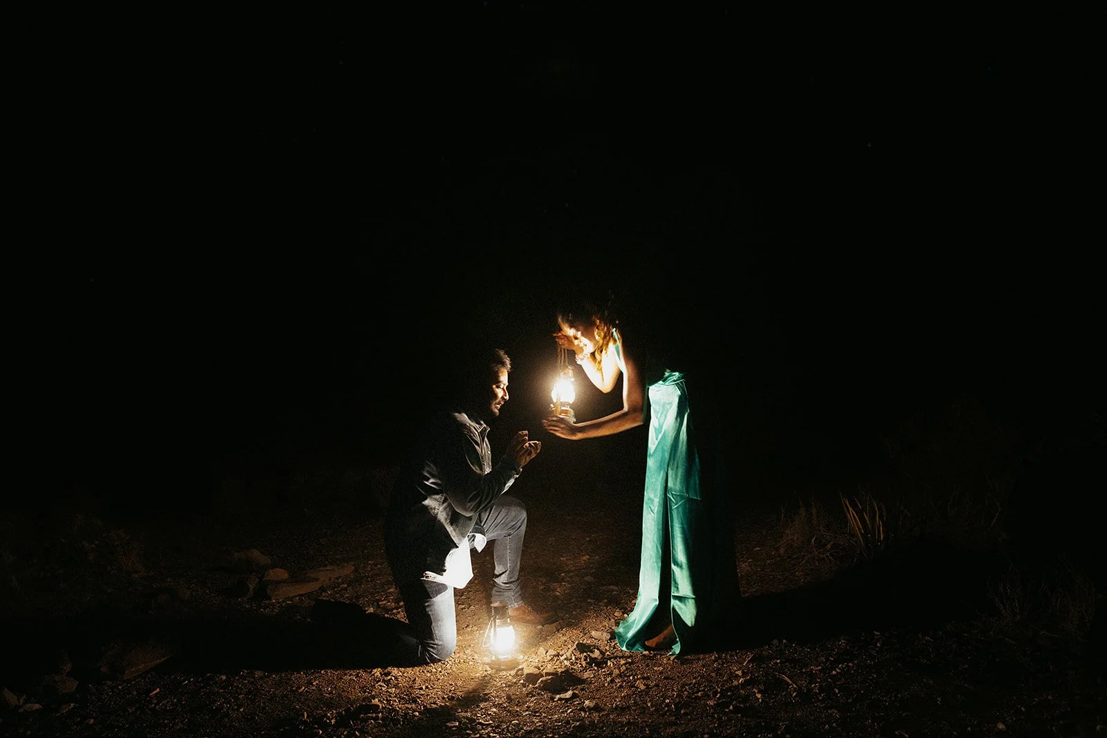 A man kneeling and woman standing in the dark, holding lanterns that illuminate their faces, with a rocky ground.