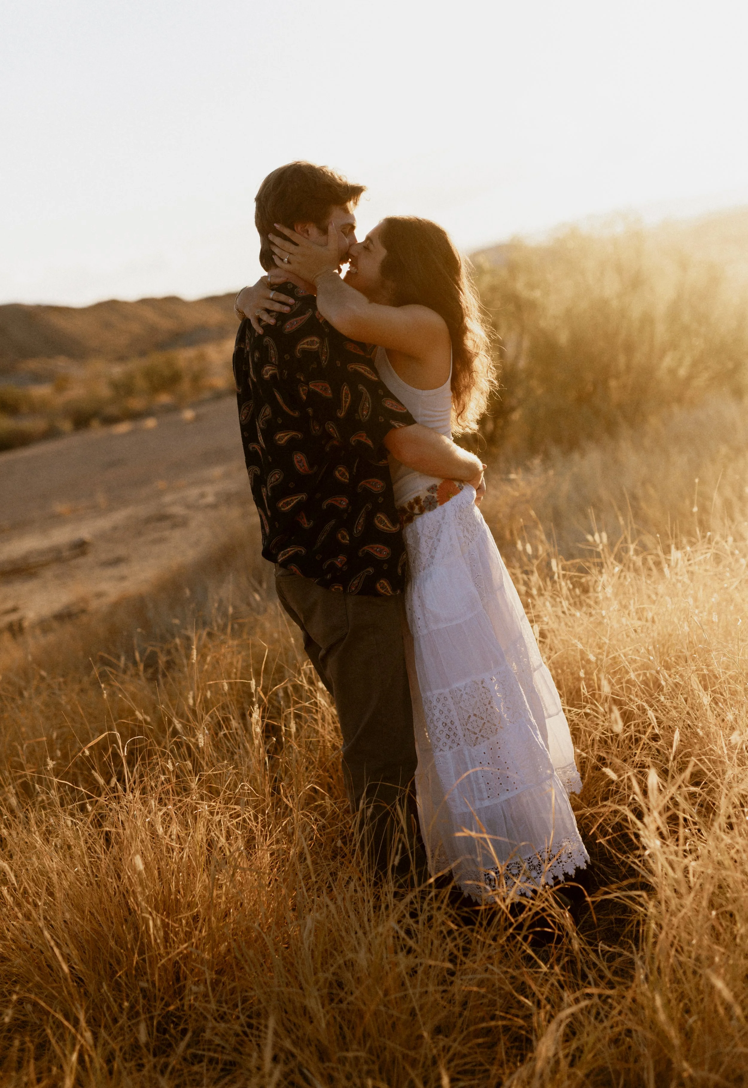 A couple embracing and kissing in a field of tall, golden grass during sunset.