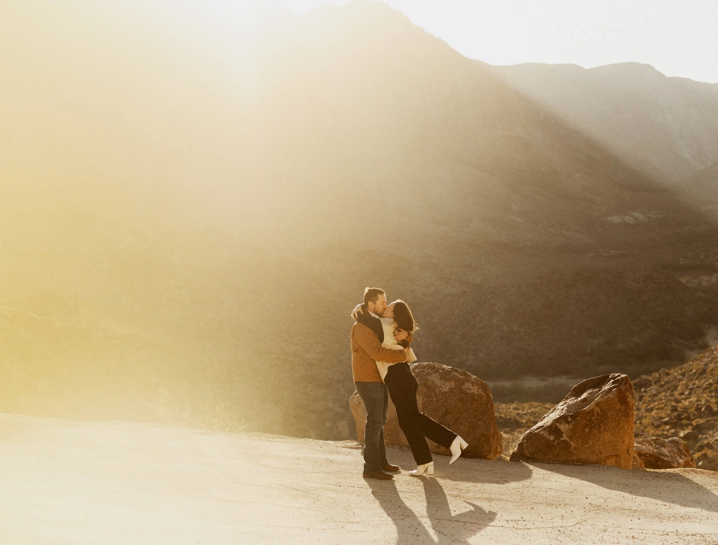 A couple embracing and kissing outdoors in front of large rocks with mountains in the background and warm sunlight.