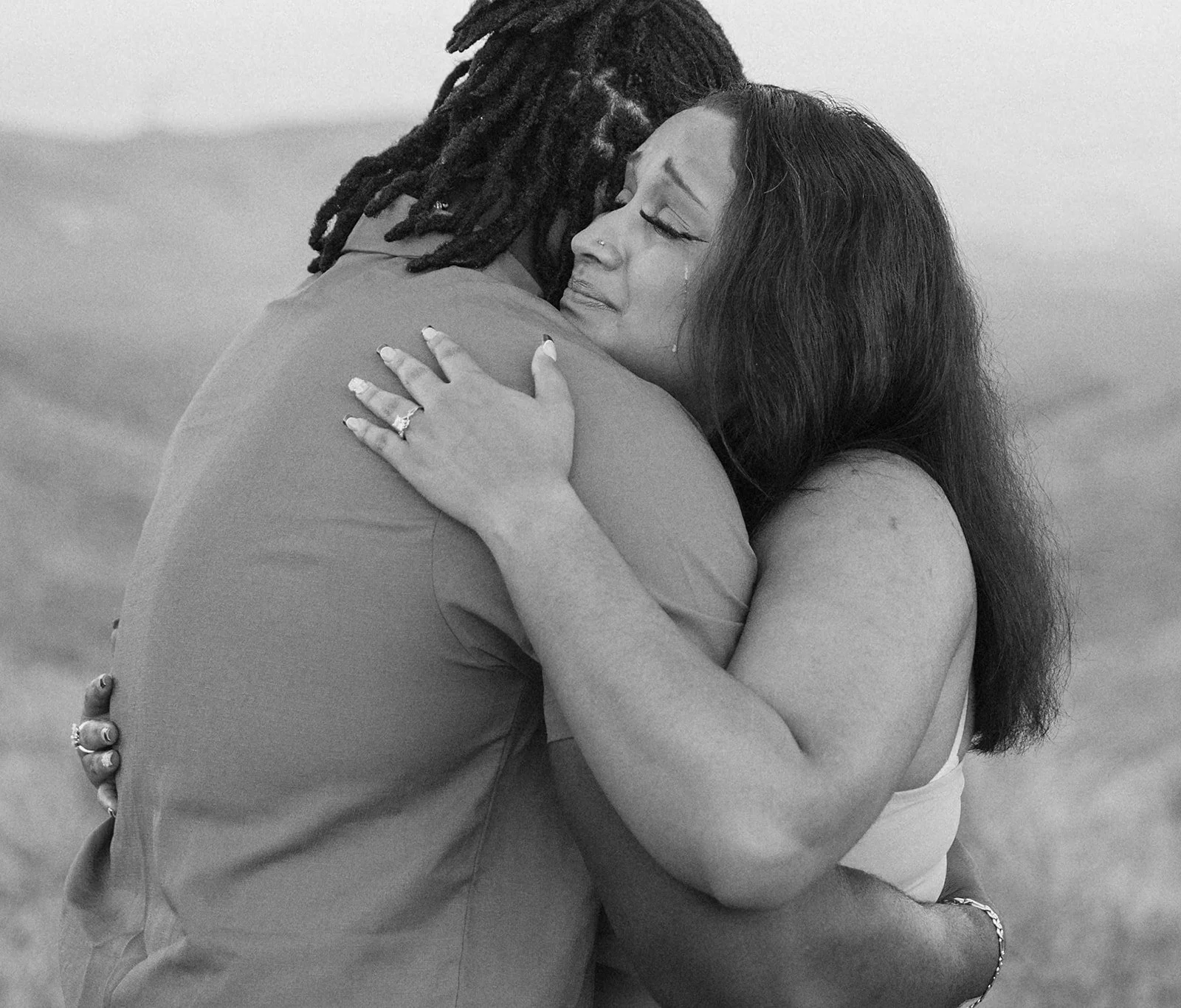 A black and white photo of a woman and man embracing tightly, with tears in her eyes.
