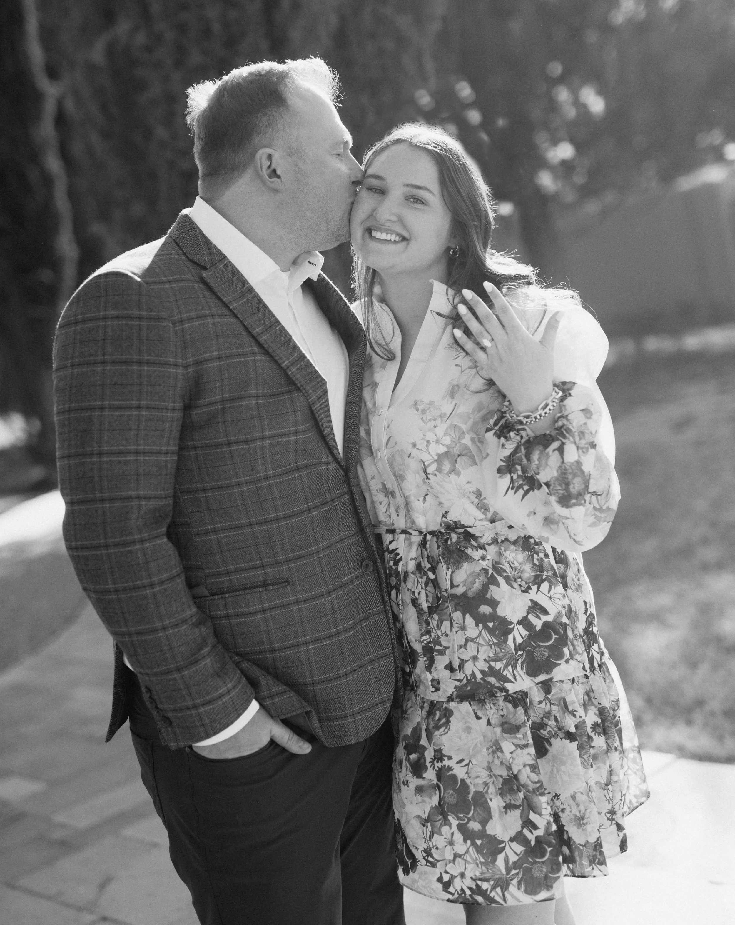 A man in a plaid suit is kissing a smiling woman in a floral dress on the cheek outdoors during daylight.