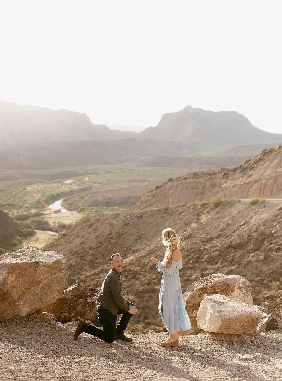 A man proposing marriage to a woman in a desert landscape with mountains in the background. The man is kneeling, and the woman is standing, wearing a light blue dress. The scene is sunlit with large rocks around them.