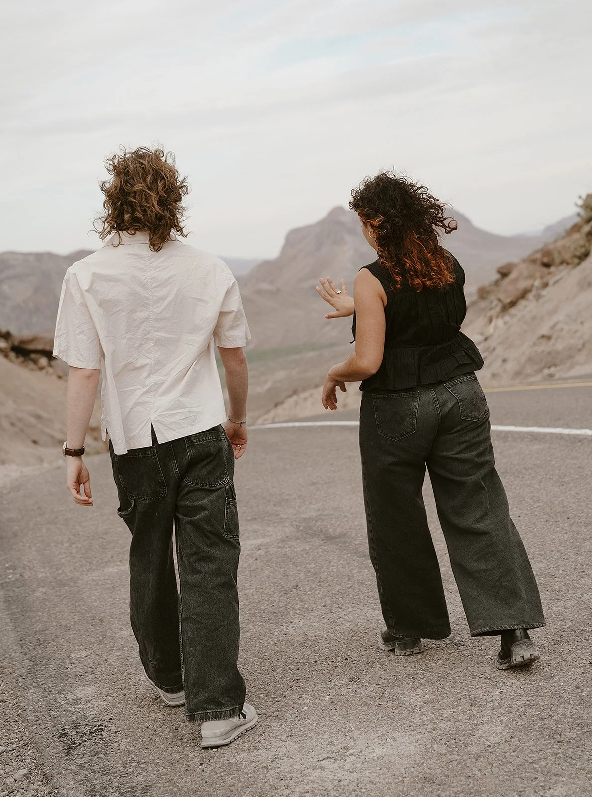 Two people walking on a paved mountain road with rocky terrain and mountains in the background, under a cloudy sky.