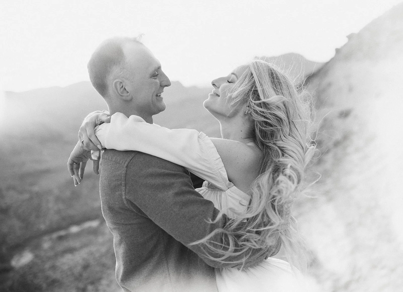 Black and white photo of a couple, man in a suit holding woman with long wavy hair, outdoors with mountains in the background.