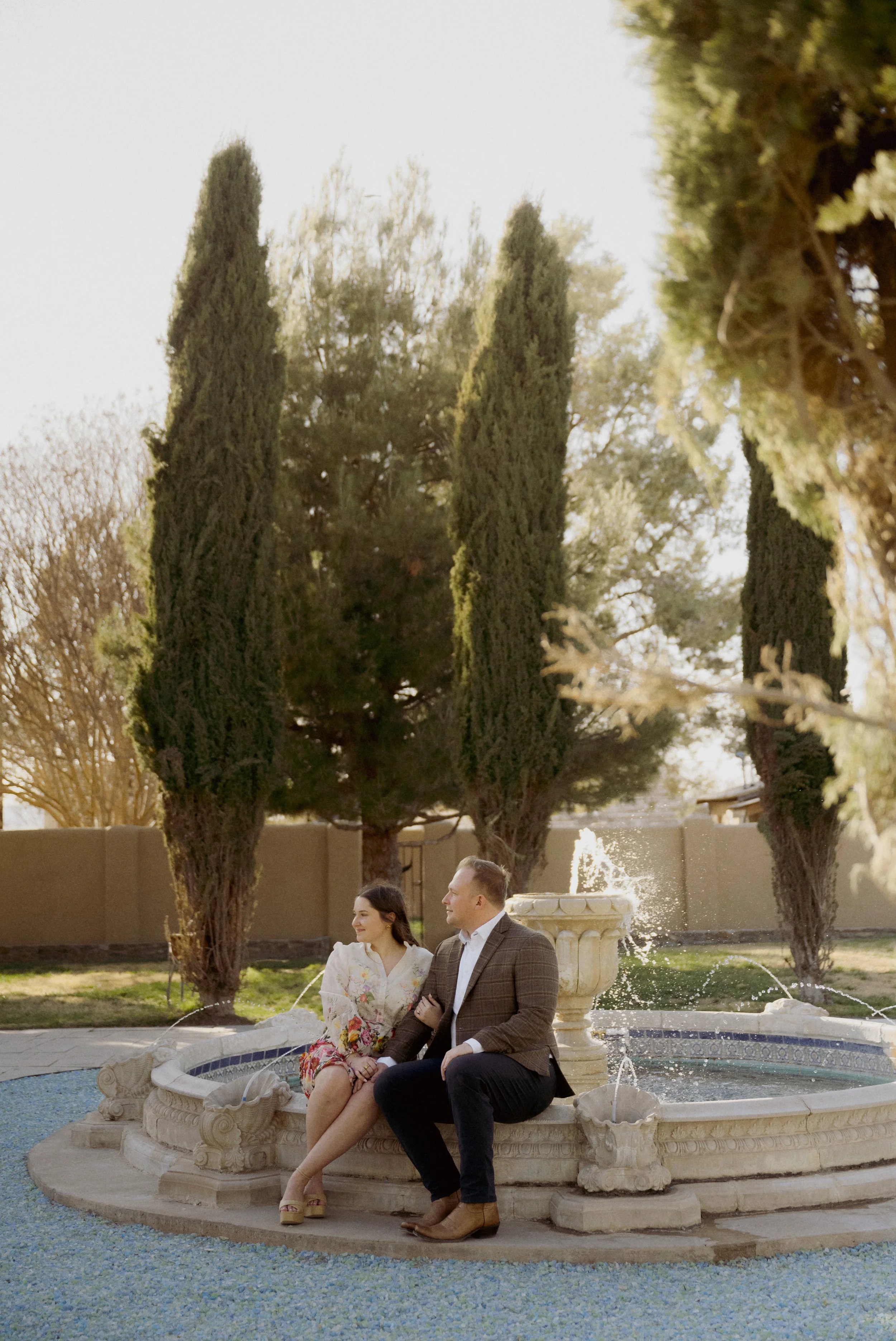 A couple sitting on a fountain in a park with tall trees behind them, during daytime.