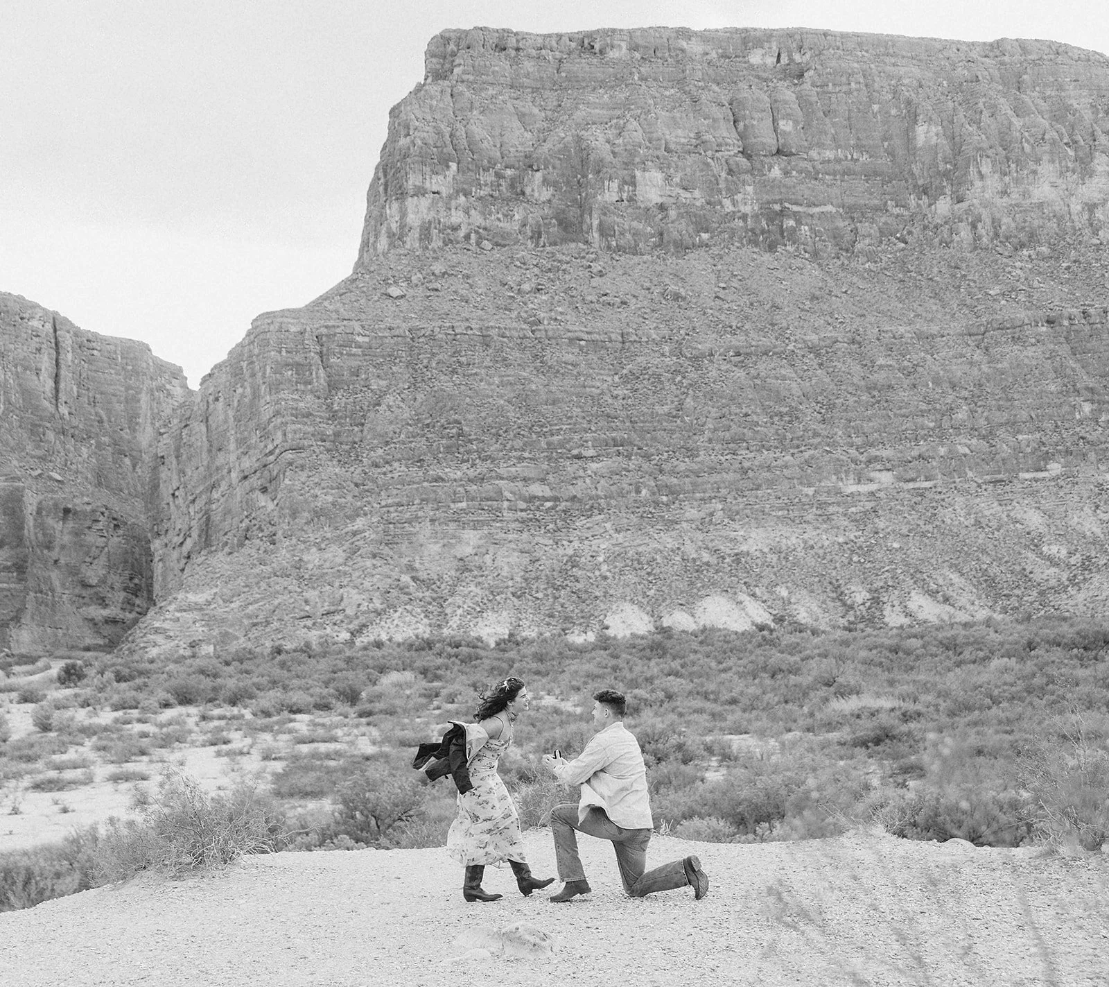 A man kneeling on one knee proposing marriage to a woman with a large mountain in the background, on a desert landscape.