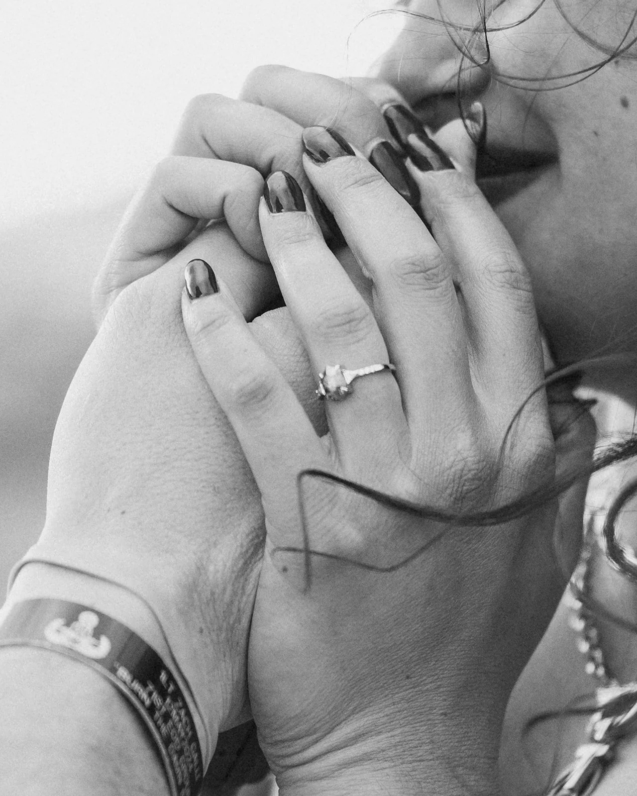 Close-up black and white photo of two hands, one with a wedding ring, gently holding each other, with part of a face visible in the background.