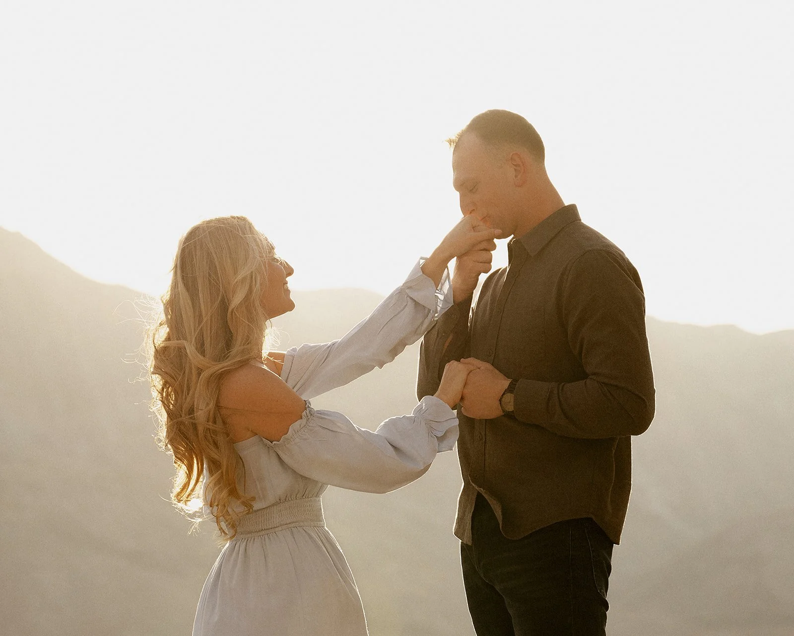 A woman giving a man a kiss on the lips outdoors at sunset, with mountains in the background.