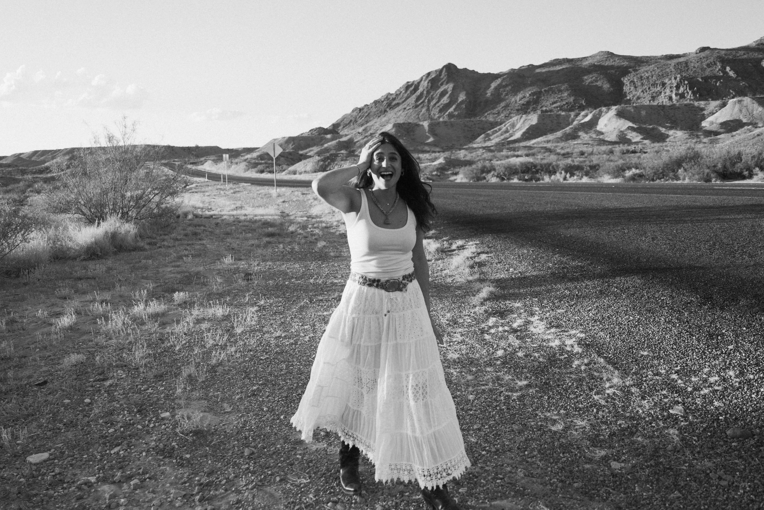 A woman with long hair, wearing a white tank top and a long, white, lace skirt, stands on a gravel roadside in a desert landscape with mountains in the background. She is smiling and holding her hand to her head.