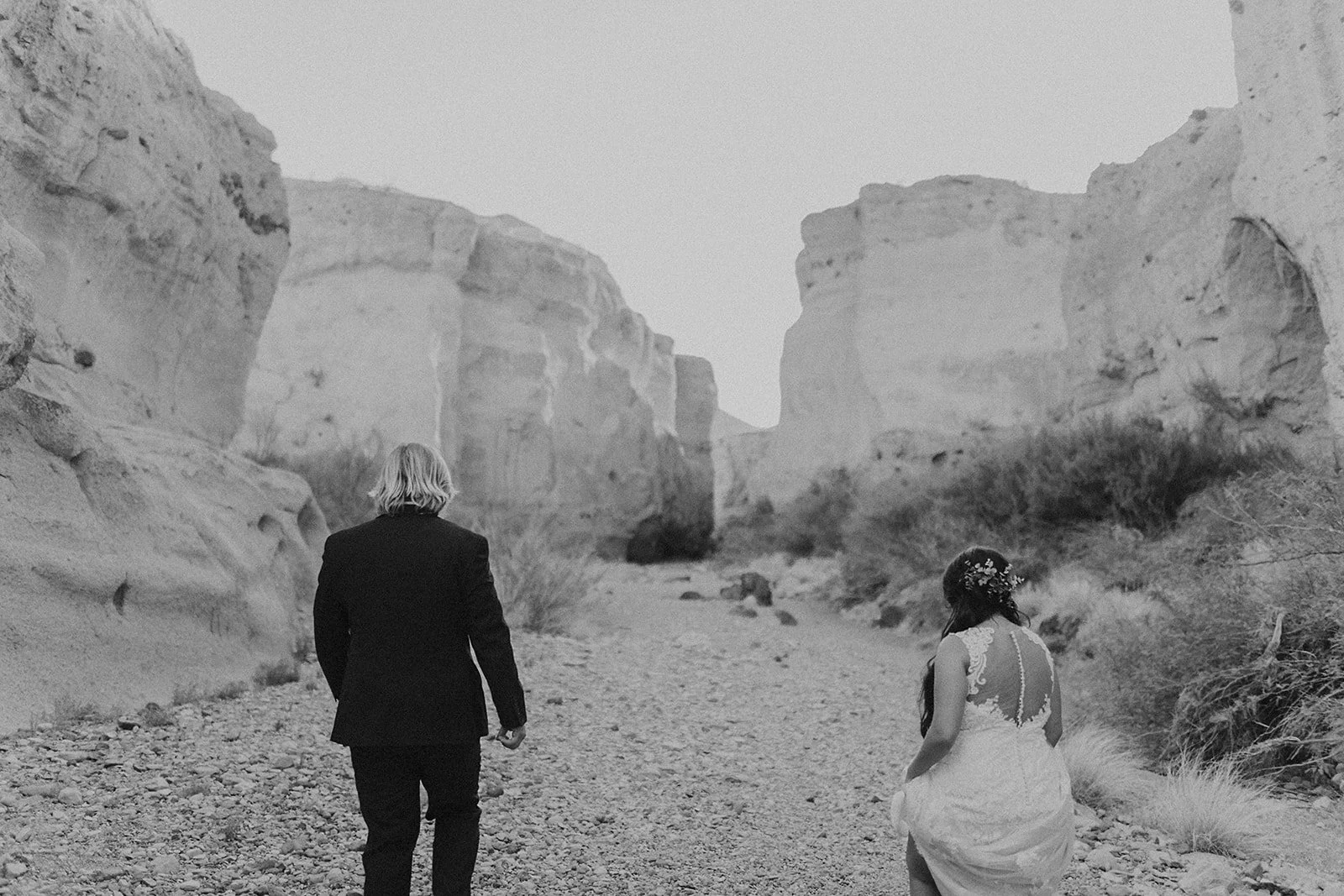 A black and white photo of a man in a suit and a woman in a wedding dress walking through a rocky canyon.