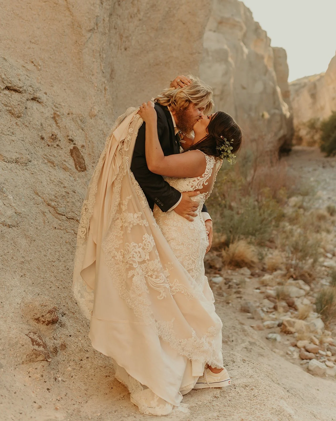 A newlywed couple sharing a kiss in a desert landscape with rocky cliffs and sparse bushes.