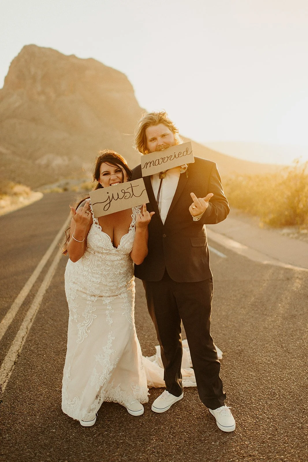 A bride and groom standing on a desert road at sunset, holding signs that read "just married" and making playful gestures.