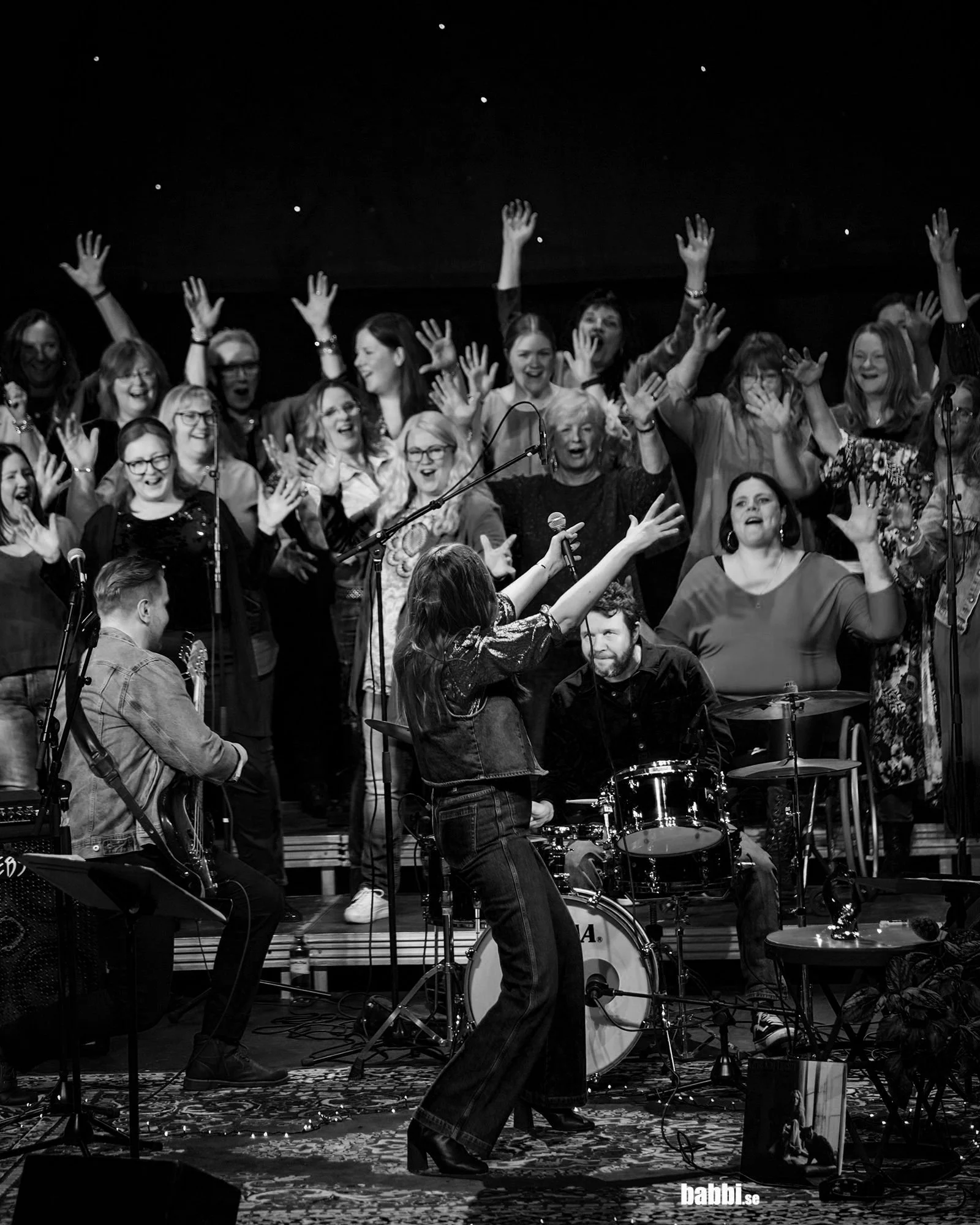 Black-and-white concert photo of a singer performing with a live band and joyful choir raising their hands onstage.