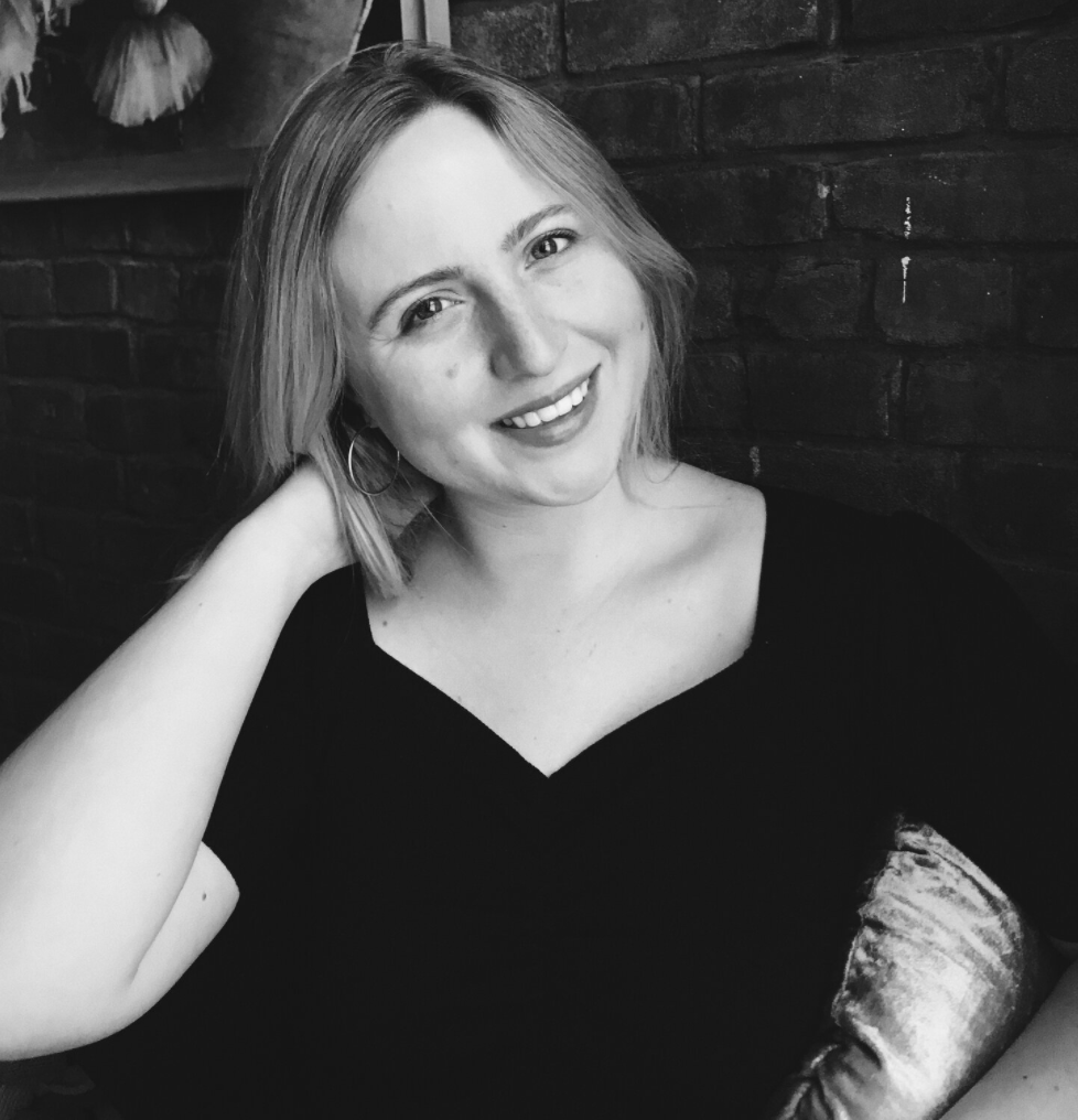 A smiling young woman with short blonde hair, wearing a black top, sitting against a brick wall, with her arm resting on the back of her head.