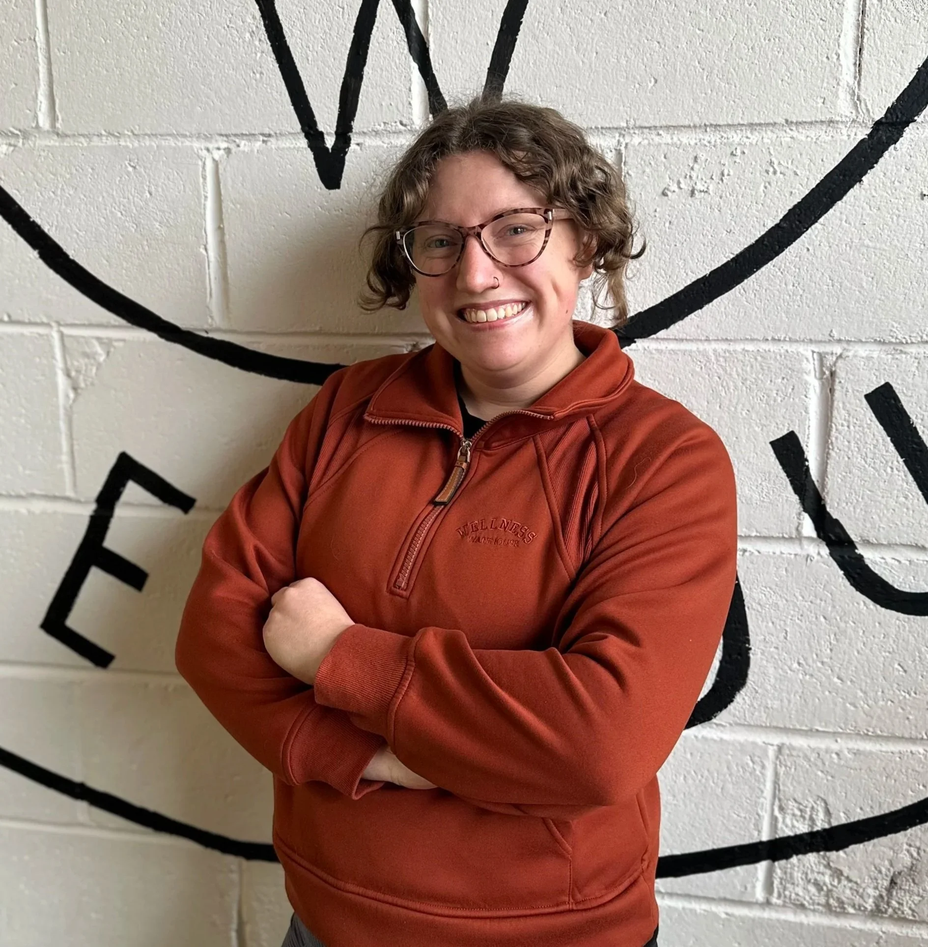 A woman with short curly hair and glasses, wearing a burnt orange zip-up jacket, smiling with arms crossed, standing in front of a white brick wall with black painted words.