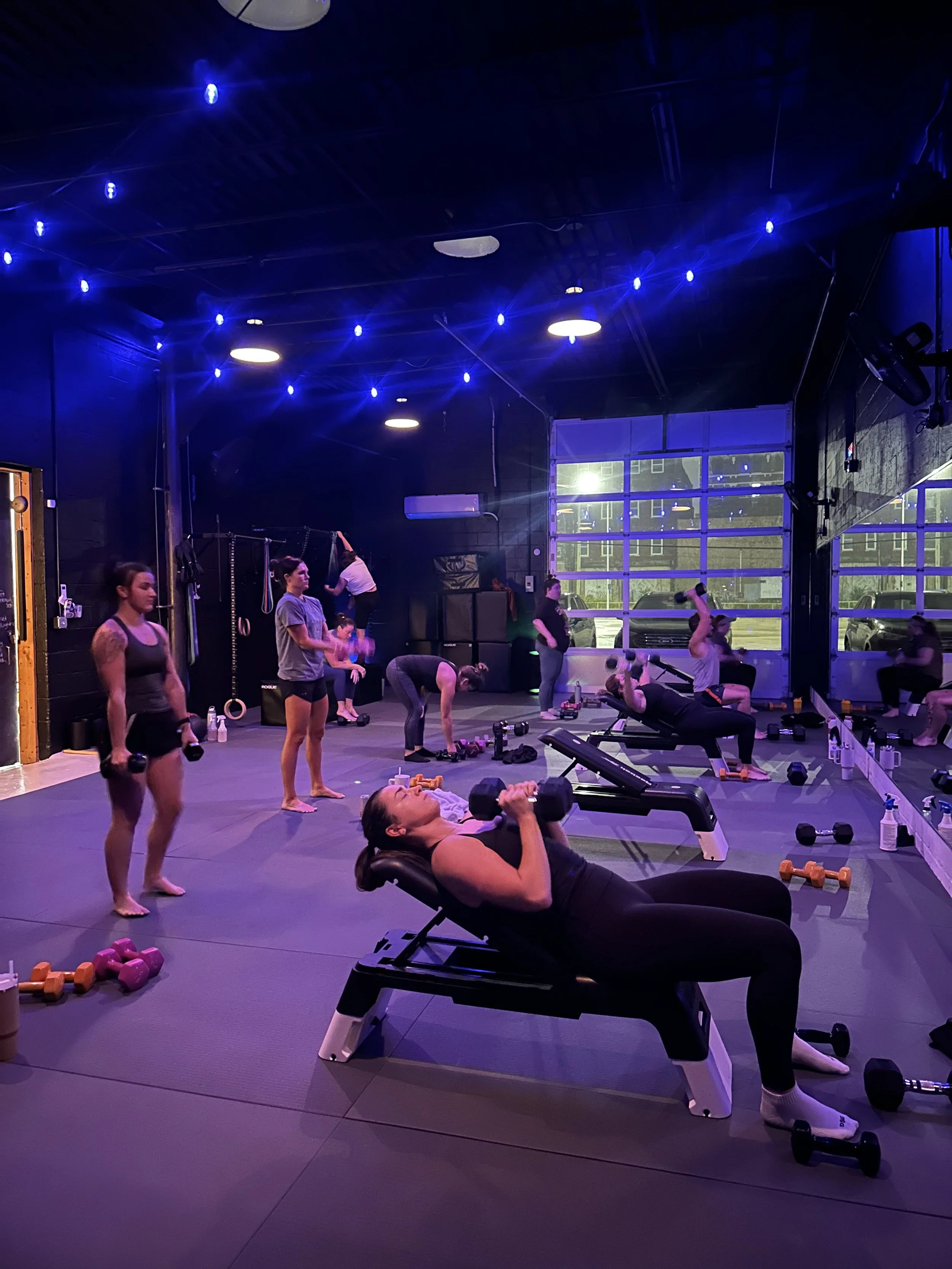 Women working out with dumbbells in a gym under blue lighting with large windows and mirrors.