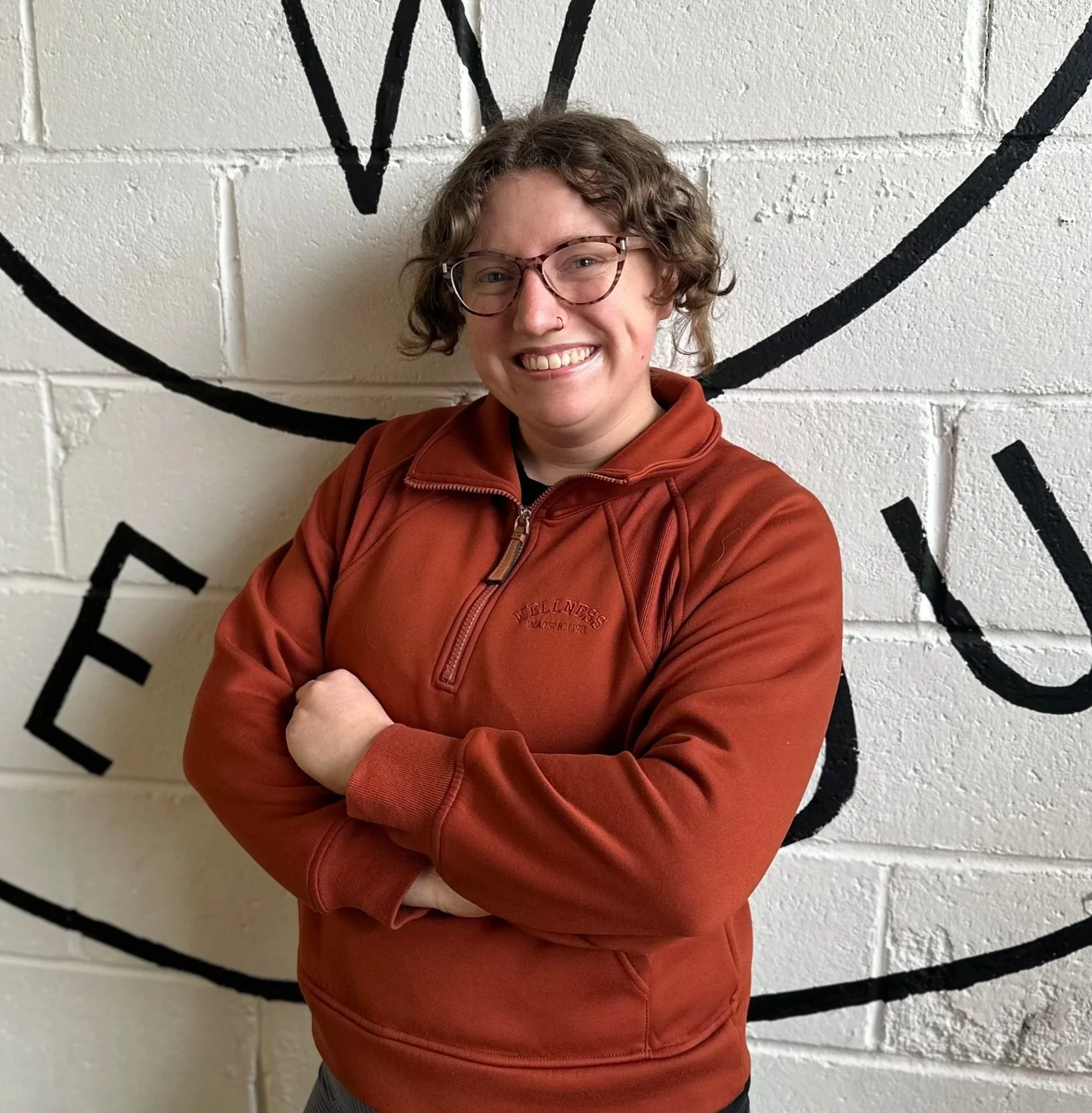 A smiling person with short curly hair and glasses, wearing a burnt orange zip-up jacket, standing with arms crossed in front of a white brick wall with black writing.