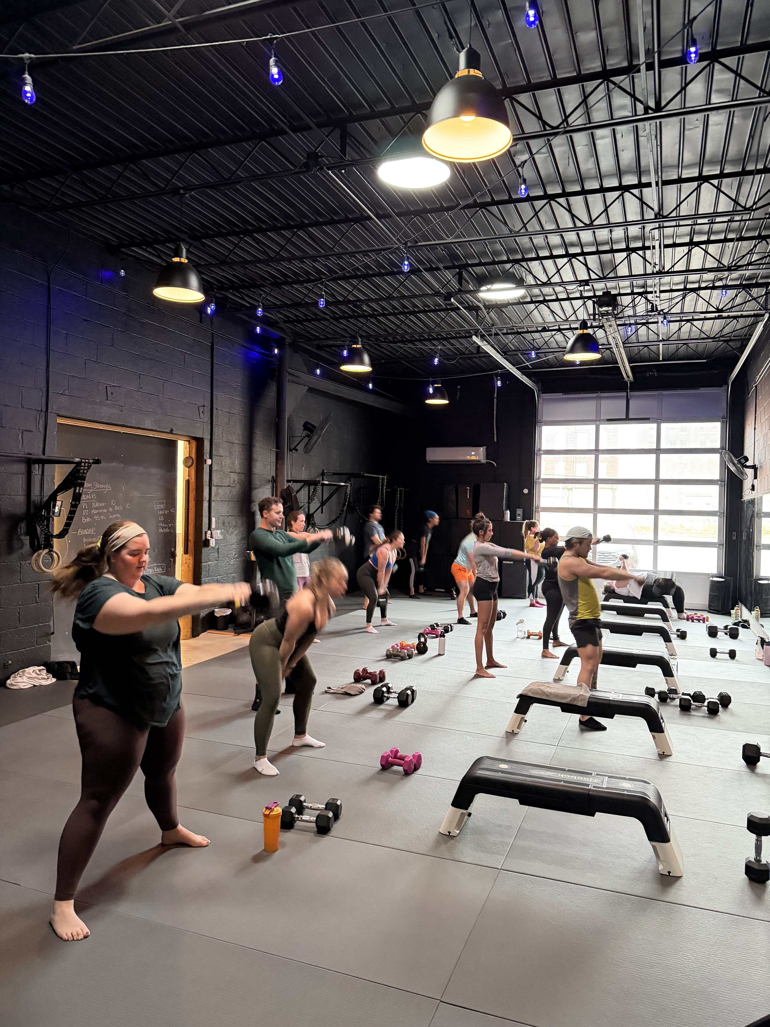 Group of people participating in a fitness class in a gym, lifting weights and doing cardio exercises, with equipment like dumbbells and benches on the floor, and large windows allowing natural light.