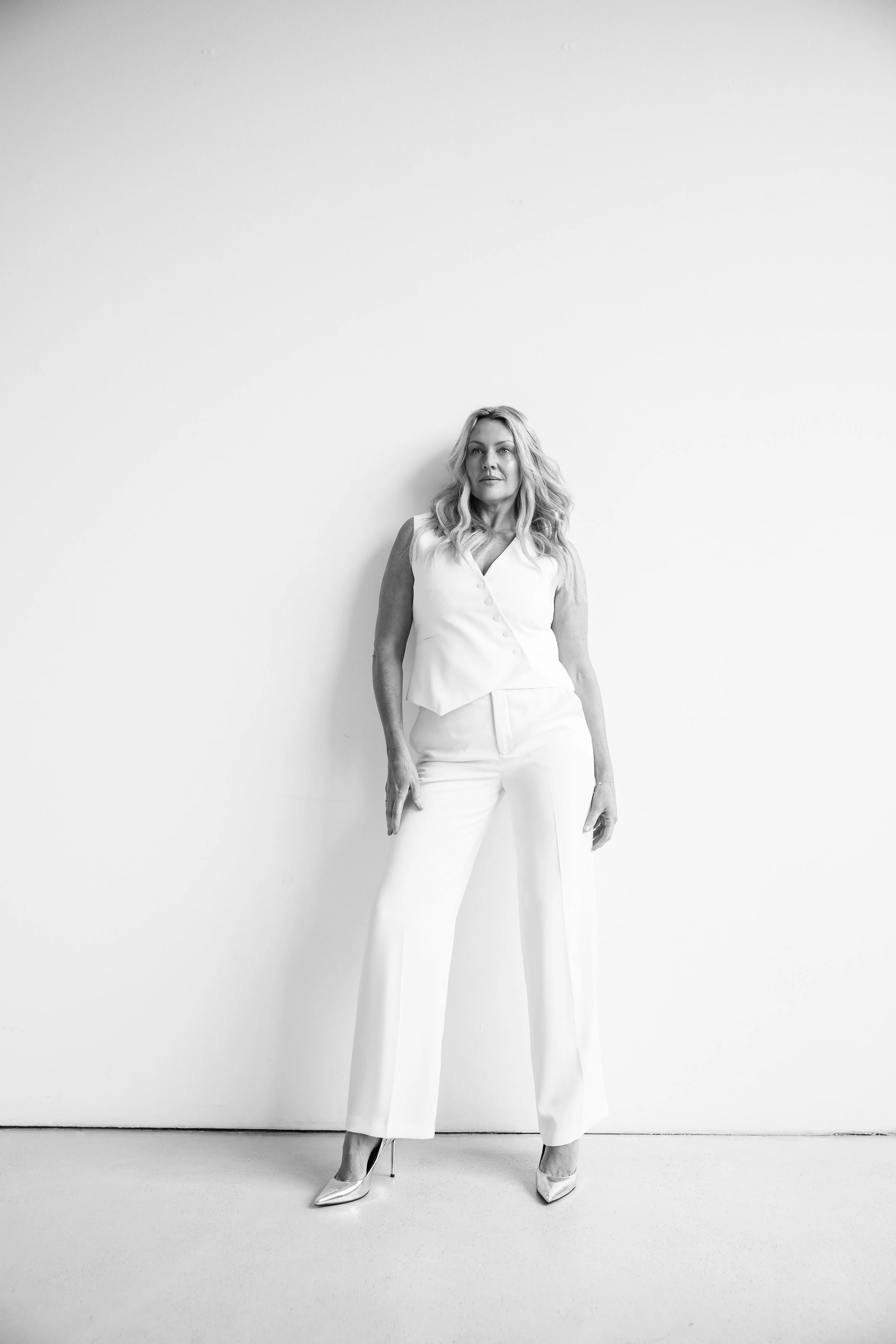 Black and white photo of a woman with long, wavy hair standing against a plain white wall. She wears a sleeveless top, wide-legged pants, and high heels, with a confident pose.