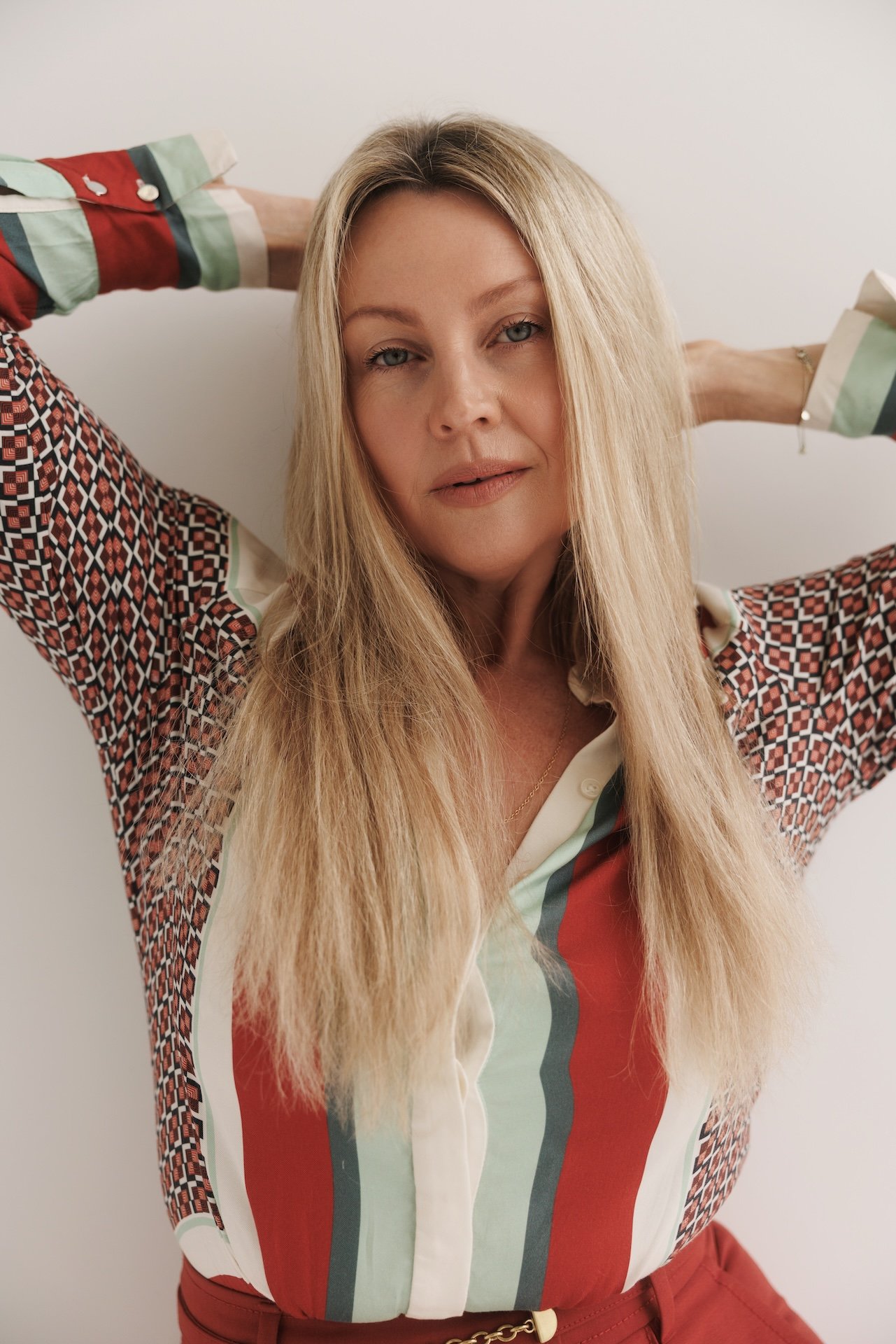 Blonde woman posing with her arms raised, wearing a colorful geometric-patterned blouse and red pants.