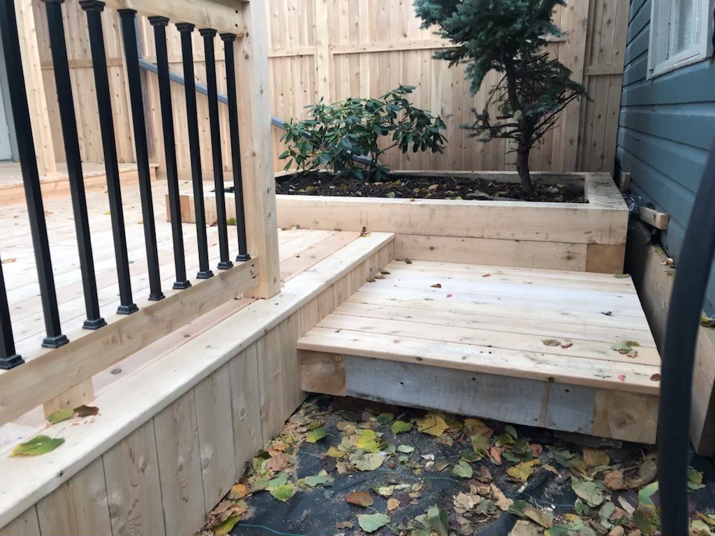 Newly built wooden deck with steps and a raised planter box with plants, surrounded by a wooden fence and blue house siding, with fallen leaves on the ground.