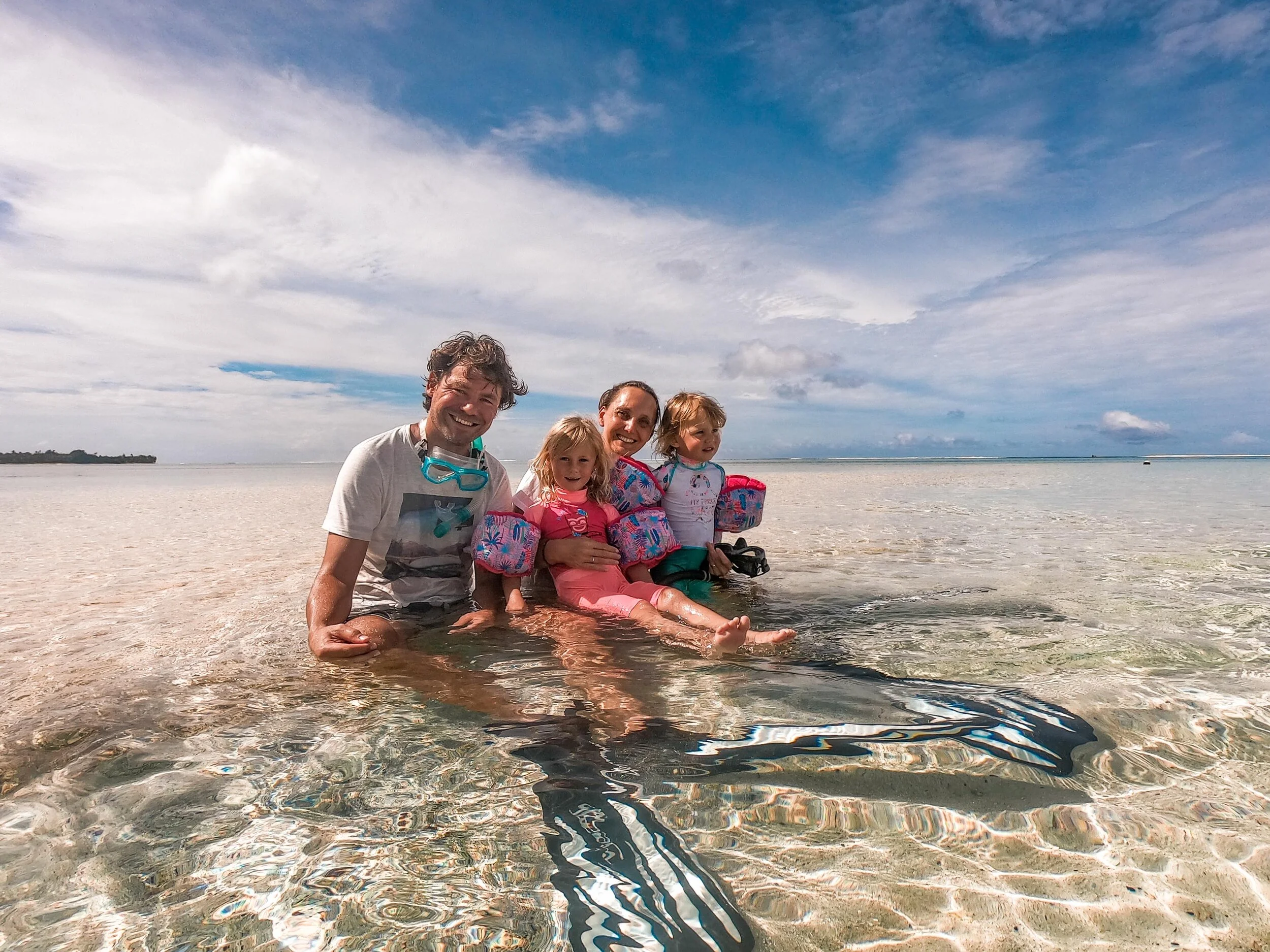 A happy family of five enjoying a day at the beach, with children and adults sitting in shallow clear water under a partly cloudy sky in Rarotonga